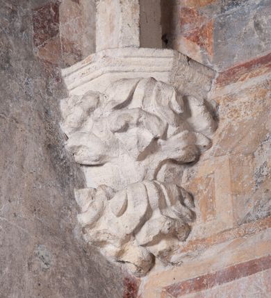 Zdjęcie nr 18: Zachowana dekoracja malarska znajduje się w tzw. starej zakrystii, określanej także jako oratorium królewskie. Dobudowano ją od północy do prezbiterium niepołomickiego kościoła. Jest to niewielka budowla na rzucie wydłużonego prostokąta, nakryta trójprzęsłowym krzyżowo-żebrowym sklepieniem, od wschodu zamknięta dwubocznie. Od zachodu w narożniku południowym widoczne jest załamanie muru, tworzącego dwie ściany wieżyczki schodowej. Do jej wnętrza prowadzi ostrołukowy portal. Oratorium oświetlają trzy ostrołukowe okna. Tylko czwarte, obecnie zamurowane, przeprute w murze północno-wschodnim pochodzi z czasów budowy kościoła. Stara zakrystia połączona jest z kościołem za pomocą ostrołukowego, profilowanego kamiennego portalu umieszczonego w środkowym przęśle ściany południowej. Na sklepieniu znajdują się rzeźbione zworniki z przedstawieniem w przęśle wschodnim głowy Chrystusa w nimbie krzyżowym, w środkowym maski liściastej, a w zachodnim motywów roślinnych. Podobnie rzeźbione motywy roślinne  pokrywają wsporniki, na które spływają żebra. 
Zachowana polichromia tworzy z architekturą starej zakrystii harmonijną całość. Białe wysklepki sklepień otoczone są wielobarwnymi, namalowanymi wzdłuż żeber, iluzjonistyczno-geometrycznymi pasami imitującymi kamienne inkrustacje w typie &quot;arte cosmatesca&quot;. Pierwotnie polichromowane były także same żebra oraz rzeźbione zworniki i wsporniki. Pionowym rytmom barwnych pasów przeciwstawiają się na ścianie wschodniej, południowej i zachodniej dwa poziome, iluzjonistyczne malowane fryzy. Górny, w odcieniach brązów jest szerszy i przypomina gzyms. Przebiega w połowie wysokości ścian, na poziomie wsporników. Poniżej znajduje się drugi, o połowę węższy, skomponowany z wielobarwnych geometrycznych rozet w rombowych polach. Fryzy wyznaczają trzy strefy dekoracji. Dolny pas uległ zniszczeniu za wyjątkiem niewielkiego fragmentu iluzjonistycznie malowanej konsolki na ścianie południowej. Dla strefy środkowej głównej tło stanowi niebieska barwa. Niestety oba te pasy obecnie są mocno uszkodzone. 
Pierwotnie pas środkowy ścian zajmowały naturalnej wielkości postacie kobiece, ujęte powyżej kolan. Z namalowanych dwunastu świętych do naszych czasów przetrwało sześć - w całości cztery (na ścianie wschodniej, południowej i zachodniej) oraz jedna bez głowy (ściana południowa) i jedna w połowie (ściana zachodnia). Są to jasno lub ciemnowłose młode kobiety, trzymające palmy męczeństwa. Ubrane są w suknie z narzuconymi płaszczami w kolorach żółtym, oliwkowym, różowym, liliowym, brązowym i błękitnym. Jedna z nich, wyobrażona ponad wejściem na klatkę schodową w wieżyczce, jest ukazana jako kobieta w podeszłym wieku z okrytą głową i dłońmi uniesionymi na wysokość piersi w geście orantki. Głowy męczennic i orantki okalają plastycznie, tłoczone w zaprawie nimby zdobione promieniami. Na niektórych głowach dziewic zachowały się ślady namalowanych brązową farbą koron o niskich sterczynach w kształcie heraldycznych lilii. 
W ścianie północno-wschodniej dwubocznego zamknięcia znajduje się obecnie zamurowane lancetowate, ostrołukowe okno z fragmentami polichromii w glifie okiennym. Pierwotnie tworzyło ją dziesięć czterolistnych obramowań wpisanych w romby, umieszczonych jeden nad drugim. Przedstawiono w nich w półpostaci aniołów z głowami otoczonymi nimbami. W dolnej części okno do  poziomu gotyckich wsporników otacza iluzjonistycznie malowana dekoracja utworzona z motywów kamiennych płyt.  
W górnej strefie oratorium wyobrażono cykl hagiograficzny, składający się z sześciu scen namalowanych w ostrołukowych polach, wyznaczonych łukami tarczowymi sklepień i ujętych w obramienia z plastycznie malowanych zębów. Ograniczający je od dołu, iluzjonistycznie malowany, jakby wystający gzyms nadaje przedstawieniom teatralną głębie. 
Na ścianie południowo-wschodniej przedstawiono bliżej niezidentyfikowaną scenę męczeństwa. Na tle ochrowo-żółtej architektury ukazano z lewej strony na pierwszym planie grupę  rycerzy, z których dwóch widocznych jest w całej postaci w średniowiecznych zbrojach stylizowanych na rzymskie, w hełmach i z ostrołukowymi tarczami trzymanymi w lewych rękach. Ich odzież ma kolor błękitny, granatowy, czerwony, fioletowy, cynobrowy. Rycerze lekko unoszą głowy w stronę półpostaci wychylającej się z okna loggi w górnej części architektury. Był to zapewne władca, dziś jego postać zachowała się tylko fragmentarycznie. Postać z okna wskazuje palcem na centralnie ukazany kocioł zawieszony pod arkadą, stanowiącą część architektury tła. Widoczna w nim jest w półpostaci jasnowłosa kobieta w białej sukni na ramiączkach z modlitewnie złożonymi rękami na piersi. Jej głowę otacza nimb. Kocioł wisi na haku za półokrągły uchwyt, pod nim pali się ognisko. Z prawej strony kotła widoczna jest fragmentarycznie zachowana postać rycerza w zbroi, który oburącz długim kijem miesza w kotle. 
Na prawo od sceny męczeństwa na ścianie południowej namalowano scenę określaną jako &quot;przed władcą&quot;. W centrum przedstawienia na tle architektury wyobrażono tron z zasiadającym na nim władcą. Jest to brodaty mężczyzna ukazany z profilu z koroną na głowie, który w prawej dłoni unosi berło skierowane w stronę przyprowadzonej do niego kobiety. Tron i architektoniczne jego obramienie malowane jest w kolorach brązu i ochry. Jego zaplecek dekoruje zawieszona pasiasta tkanina. Ciemnowłosy władca ubrany jest w turkusową szatę z narzuconym na ramiona brązowym płaszczem. Za tronem z lewej strony stoi paź o kędzierzawej fryzurze wsparty na trzymanym ostrzem w dół mieczu. Stojąca przed władcą kobieta ubrana jest w czerwoną długą suknię. Ma jasne włosy, a jej głowę okala nimb. Ręce ma skrzyżowane i związane na wysokości pasa. Obok niej stoi inna postać także w długiej szacie, z uniesioną prawą dłonią. Za świętą widoczne są inne postacie, niestety skala zniszczenia malowidła uniemożliwia ich opis i identyfikację. 
Trzecie malowidło, usytuowane nad wejściem do oratorium, ukazuje scenę dysputyt. W centrum przedstawienia pod architektonicznym zadaszeniem w formie portyku lub altany o kasetonowym stropie, wspartym na dwóch czworobocznych filarach, ukazano widoczne z profilu trzy siedzące  kobiety. Jedna z lewej strony a dwie naprzeciwko z prawej. Kobieta z lewej  ma odkryte jasne, długie włosy. Jej głowę otacza nimb. Na wysokość piersi unosi obie dłonie, sprawiając wrażenie, jakby w nich trzymała owalny przedmiot. Ubrana jest w ciemnoczerwoną długą suknię, zakrywającą nogi. Naprzeciw niej siedzą dwie inne niewiasty o długich, jasnych włosach w prostych koronach na głowach. Jedna z nich unosi swoją prawą rękę. Obie ubrane są w długie suknie w kolorze ochry. Za nimi pomiędzy filarami widoczna jest rozpięta zasłona w pionowe oliwkowe, zielone, białe i czerwone pasy. Na podłodze leży dywan dekorowany w żółte, zielone, niebieskie i czerwone pasy. 
Mocno zniszczone w dolnej części czwarte przedstawienie ilustruje scenę chłosty. Z lewej strony kompozycji wyobrażono budowlę z architektoniczną loggią, z której w półpostaci wychyla się władca. W prawej ręce poziomo trzyma berło skierowane w stronę grupy osób, za którymi wykonywana jest kara chłosty. Władca, podobnie jak w drugiej scenie, został ukazany jako mężczyzna o ciemnym zaroście i takich samych włosach. Na głowie ma koronę. Ubrany jest w brązowe szaty o różnych odcieniach. Centralnie przedstawioną stłoczoną grupę tworzy czterech żołnierzy oraz starszy mężczyzna w zawoju na głowie z siwą, długą brodą. Żołnierze mają zbroje i hełmy. Jeden z nich, najbliżej stojący z lewej przedstawiony został w profilu z głową i prawą ręką skierowaną w stronę króla. Także w stronę władcy zwrócony jest mężczyzna w zawoju na głowie z dwiema dłońmi uniesionymi na wysokość piersi. Z prawej strony kompozycji namalowano, dziś fragmentarycznie zachowany, rodzaj drewnianej szubienicy, utworzonej z poprzecznie zawieszonego na drewnianych podporach drążka. Nad dróżką do góry nogami wisi postać (dziś zniszczona), z której zachowały się jedynie stopy związane w kostkach sznurem. W jej stronę zwraca się jeden z żołnierzy ze środkowej grupy. Po prawej stronie szubienicy widoczna jest część postaci oprawcy, z której zachował się tylko fragment głowy oraz uniesiona lewa ręka z drewnianą pałką. 
Dwie ostatnie sceny umieszczono na zachodnim murze oratorium. Pierwsza z nich przedstawia strącenie bożka i została połączona z drugą ukazującą uwolnienia z więzienia. Część centralną kompozycji stanowi potrzaskana, wywracająca się kolumna, z której spada szatan w postaci żywego demona pokrytego sierścią. Strącił go widoczny w półpostaci w górnej części malowidła anioł trzymający w prawej ręce miecz. Z lewej strony kompozycji  klęczy na skalistej ziemi święta ubrana w długą ciemnoczerwoną suknię. Niestety nie zachowała się górna część jej postaci. Za nią stoi stłoczona grupa przerażonych świadków tego wydarzenia. Natomiast z prawej strony malowidła wyobrażono scenę uwolnienia z więzienia. Jej zasadniczym elementem jest architektoniczna struktura nawiązująca kształtem do wieży mieszkalnej, więzienia bądź bramy miejskiej. Święta ze związanymi rękami w ciemnoczerwonej sukni wychodzi z niej z prawej strony. W centrum budowli widoczne jest półokrągłe okno, a w nim zapewne jej popiersie o długich jasnych włosach, trzymające w dłoniach jakiś przedmiot. Wszystkie sceny cyklu w jedną całość wiąże gładkie niebieskie tło. Niezachowana kompozycja polichromii na ścianie północnej musiała odbiegać swym układem od pozostałych ścian. Płaszczyzna ścian została podzielona za pomocą węższego dolnego fryzu na dwie strefy. Górną część najprawdopodobniej zajmowały duże postacie stojące (fragment jednej z nich zachował się w sąsiedztwie okna w ścianie północno-wschodniej). Pod fryzem w części dolnej najprawdopodobniej była namalowana iluzjonistycznie wisząca kotara.   
    




