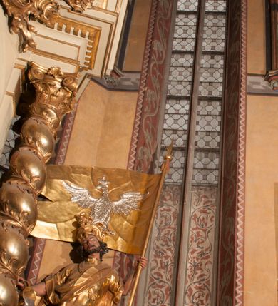 Zdjęcie nr 9: Malowidła z przedstawieniem św. Michała Archanioła pokonującego szatana i św. Jerzego walczącego ze smokiem zostały wykonane na ścianie północnej w dwóch wnękach podokiennych. Obie kompozycje powstały w polach o kształcie wydłużonego prostokąta. Święty Michał Archanioł został przedstawiony frontalnie w całej postaci z głową w ujęciu trzech czwartych w lewo. W uniesionej ponad głowę prawej ręce trzyma włócznię, wbitą w deptanego pod nogami szatana, którą jednocześnie podtrzymuje lewą ręką. Z tyłu widać jego rozpostarte skrzydła, skrzyżowane nad głową. Archanioł ma owalną twarz o wydatnym nosie oraz jasne włosy spływające na ramiona. Ubrany jest w przepasaną  tunikę sięgającą goleni i w narzucony na ramiona płaszcz. Postać diabła pod jego stopami obutymi w buty jest prawie niewidoczna. W podobny sposób przedstawiono św. Jerzego. Święty został ukazany frontalnie na rozstawionych nogach w całej postaci ujętej w kontrapoście z głową w trzech czwartych skierowaną w lewo. Prawą rękę ma uniesioną ponad głową i zgiętą w łokciu. Trzyma w niej lancę podtrzymywaną lewą ręką. Pod jego stopami widoczny jest zarys zwiniętego smoka o zarysie bazyliszka. Św. Jerzy ubrany jest w strój rycerski. Na głowie ma spiczasto zakończony hełm z kolczugą. Ciało okrywa mocno taliowana w pasie tunika zdobiona u dołu ornamentem z motywem trójkąta równoramiennego. Na nogach ma założone wysokie buty. W obu przedstawieniach widoczne są ciemne ugry, ciemna czerwień, żółcień, błękit i brąz. Tła są szare. 