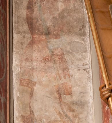 Zdjęcie nr 3: Malowidła z przedstawieniem św. Michała Archanioła pokonującego szatana i św. Jerzego walczącego ze smokiem zostały wykonane na ścianie północnej w dwóch wnękach podokiennych. Obie kompozycje powstały w polach o kształcie wydłużonego prostokąta. Święty Michał Archanioł został przedstawiony frontalnie w całej postaci z głową w ujęciu trzech czwartych w lewo. W uniesionej ponad głowę prawej ręce trzyma włócznię, wbitą w deptanego pod nogami szatana, którą jednocześnie podtrzymuje lewą ręką. Z tyłu widać jego rozpostarte skrzydła, skrzyżowane nad głową. Archanioł ma owalną twarz o wydatnym nosie oraz jasne włosy spływające na ramiona. Ubrany jest w przepasaną  tunikę sięgającą goleni i w narzucony na ramiona płaszcz. Postać diabła pod jego stopami obutymi w buty jest prawie niewidoczna. W podobny sposób przedstawiono św. Jerzego. Święty został ukazany frontalnie na rozstawionych nogach w całej postaci ujętej w kontrapoście z głową w trzech czwartych skierowaną w lewo. Prawą rękę ma uniesioną ponad głową i zgiętą w łokciu. Trzyma w niej lancę podtrzymywaną lewą ręką. Pod jego stopami widoczny jest zarys zwiniętego smoka o zarysie bazyliszka. Św. Jerzy ubrany jest w strój rycerski. Na głowie ma spiczasto zakończony hełm z kolczugą. Ciało okrywa mocno taliowana w pasie tunika zdobiona u dołu ornamentem z motywem trójkąta równoramiennego. Na nogach ma założone wysokie buty. W obu przedstawieniach widoczne są ciemne ugry, ciemna czerwień, żółcień, błękit i brąz. Tła są szare. 