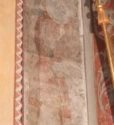 Zdjęcie nr 2: Malowidła z przedstawieniem św. Michała Archanioła pokonującego szatana i św. Jerzego walczącego ze smokiem zostały wykonane na ścianie północnej w dwóch wnękach podokiennych. Obie kompozycje powstały w polach o kształcie wydłużonego prostokąta. Święty Michał Archanioł został przedstawiony frontalnie w całej postaci z głową w ujęciu trzech czwartych w lewo. W uniesionej ponad głowę prawej ręce trzyma włócznię, wbitą w deptanego pod nogami szatana, którą jednocześnie podtrzymuje lewą ręką. Z tyłu widać jego rozpostarte skrzydła, skrzyżowane nad głową. Archanioł ma owalną twarz o wydatnym nosie oraz jasne włosy spływające na ramiona. Ubrany jest w przepasaną  tunikę sięgającą goleni i w narzucony na ramiona płaszcz. Postać diabła pod jego stopami obutymi w buty jest prawie niewidoczna. W podobny sposób przedstawiono św. Jerzego. Święty został ukazany frontalnie na rozstawionych nogach w całej postaci ujętej w kontrapoście z głową w trzech czwartych skierowaną w lewo. Prawą rękę ma uniesioną ponad głową i zgiętą w łokciu. Trzyma w niej lancę podtrzymywaną lewą ręką. Pod jego stopami widoczny jest zarys zwiniętego smoka o zarysie bazyliszka. Św. Jerzy ubrany jest w strój rycerski. Na głowie ma spiczasto zakończony hełm z kolczugą. Ciało okrywa mocno taliowana w pasie tunika zdobiona u dołu ornamentem z motywem trójkąta równoramiennego. Na nogach ma założone wysokie buty. W obu przedstawieniach widoczne są ciemne ugry, ciemna czerwień, żółcień, błękit i brąz. Tła są szare. 