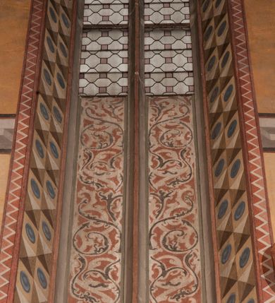 Zdjęcie nr 6: Malowidła z przedstawieniem św. Michała Archanioła pokonującego szatana i św. Jerzego walczącego ze smokiem zostały wykonane na ścianie północnej w dwóch wnękach podokiennych. Obie kompozycje powstały w polach o kształcie wydłużonego prostokąta. Święty Michał Archanioł został przedstawiony frontalnie w całej postaci z głową w ujęciu trzech czwartych w lewo. W uniesionej ponad głowę prawej ręce trzyma włócznię, wbitą w deptanego pod nogami szatana, którą jednocześnie podtrzymuje lewą ręką. Z tyłu widać jego rozpostarte skrzydła, skrzyżowane nad głową. Archanioł ma owalną twarz o wydatnym nosie oraz jasne włosy spływające na ramiona. Ubrany jest w przepasaną  tunikę sięgającą goleni i w narzucony na ramiona płaszcz. Postać diabła pod jego stopami obutymi w buty jest prawie niewidoczna. W podobny sposób przedstawiono św. Jerzego. Święty został ukazany frontalnie na rozstawionych nogach w całej postaci ujętej w kontrapoście z głową w trzech czwartych skierowaną w lewo. Prawą rękę ma uniesioną ponad głową i zgiętą w łokciu. Trzyma w niej lancę podtrzymywaną lewą ręką. Pod jego stopami widoczny jest zarys zwiniętego smoka o zarysie bazyliszka. Św. Jerzy ubrany jest w strój rycerski. Na głowie ma spiczasto zakończony hełm z kolczugą. Ciało okrywa mocno taliowana w pasie tunika zdobiona u dołu ornamentem z motywem trójkąta równoramiennego. Na nogach ma założone wysokie buty. W obu przedstawieniach widoczne są ciemne ugry, ciemna czerwień, żółcień, błękit i brąz. Tła są szare. 