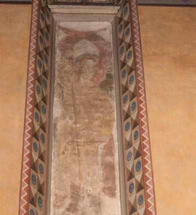 Zdjęcie nr 5: Malowidła z przedstawieniem św. Michała Archanioła pokonującego szatana i św. Jerzego walczącego ze smokiem zostały wykonane na ścianie północnej w dwóch wnękach podokiennych. Obie kompozycje powstały w polach o kształcie wydłużonego prostokąta. Święty Michał Archanioł został przedstawiony frontalnie w całej postaci z głową w ujęciu trzech czwartych w lewo. W uniesionej ponad głowę prawej ręce trzyma włócznię, wbitą w deptanego pod nogami szatana, którą jednocześnie podtrzymuje lewą ręką. Z tyłu widać jego rozpostarte skrzydła, skrzyżowane nad głową. Archanioł ma owalną twarz o wydatnym nosie oraz jasne włosy spływające na ramiona. Ubrany jest w przepasaną  tunikę sięgającą goleni i w narzucony na ramiona płaszcz. Postać diabła pod jego stopami obutymi w buty jest prawie niewidoczna. W podobny sposób przedstawiono św. Jerzego. Święty został ukazany frontalnie na rozstawionych nogach w całej postaci ujętej w kontrapoście z głową w trzech czwartych skierowaną w lewo. Prawą rękę ma uniesioną ponad głową i zgiętą w łokciu. Trzyma w niej lancę podtrzymywaną lewą ręką. Pod jego stopami widoczny jest zarys zwiniętego smoka o zarysie bazyliszka. Św. Jerzy ubrany jest w strój rycerski. Na głowie ma spiczasto zakończony hełm z kolczugą. Ciało okrywa mocno taliowana w pasie tunika zdobiona u dołu ornamentem z motywem trójkąta równoramiennego. Na nogach ma założone wysokie buty. W obu przedstawieniach widoczne są ciemne ugry, ciemna czerwień, żółcień, błękit i brąz. Tła są szare. 