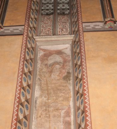 Zdjęcie nr 11: Malowidła z przedstawieniem św. Michała Archanioła pokonującego szatana i św. Jerzego walczącego ze smokiem zostały wykonane na ścianie północnej w dwóch wnękach podokiennych. Obie kompozycje powstały w polach o kształcie wydłużonego prostokąta. Święty Michał Archanioł został przedstawiony frontalnie w całej postaci z głową w ujęciu trzech czwartych w lewo. W uniesionej ponad głowę prawej ręce trzyma włócznię, wbitą w deptanego pod nogami szatana, którą jednocześnie podtrzymuje lewą ręką. Z tyłu widać jego rozpostarte skrzydła, skrzyżowane nad głową. Archanioł ma owalną twarz o wydatnym nosie oraz jasne włosy spływające na ramiona. Ubrany jest w przepasaną  tunikę sięgającą goleni i w narzucony na ramiona płaszcz. Postać diabła pod jego stopami obutymi w buty jest prawie niewidoczna. W podobny sposób przedstawiono św. Jerzego. Święty został ukazany frontalnie na rozstawionych nogach w całej postaci ujętej w kontrapoście z głową w trzech czwartych skierowaną w lewo. Prawą rękę ma uniesioną ponad głową i zgiętą w łokciu. Trzyma w niej lancę podtrzymywaną lewą ręką. Pod jego stopami widoczny jest zarys zwiniętego smoka o zarysie bazyliszka. Św. Jerzy ubrany jest w strój rycerski. Na głowie ma spiczasto zakończony hełm z kolczugą. Ciało okrywa mocno taliowana w pasie tunika zdobiona u dołu ornamentem z motywem trójkąta równoramiennego. Na nogach ma założone wysokie buty. W obu przedstawieniach widoczne są ciemne ugry, ciemna czerwień, żółcień, błękit i brąz. Tła są szare. 