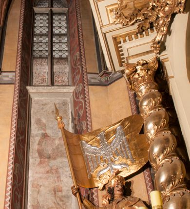 Zdjęcie nr 12: Malowidła z przedstawieniem św. Michała Archanioła pokonującego szatana i św. Jerzego walczącego ze smokiem zostały wykonane na ścianie północnej w dwóch wnękach podokiennych. Obie kompozycje powstały w polach o kształcie wydłużonego prostokąta. Święty Michał Archanioł został przedstawiony frontalnie w całej postaci z głową w ujęciu trzech czwartych w lewo. W uniesionej ponad głowę prawej ręce trzyma włócznię, wbitą w deptanego pod nogami szatana, którą jednocześnie podtrzymuje lewą ręką. Z tyłu widać jego rozpostarte skrzydła, skrzyżowane nad głową. Archanioł ma owalną twarz o wydatnym nosie oraz jasne włosy spływające na ramiona. Ubrany jest w przepasaną  tunikę sięgającą goleni i w narzucony na ramiona płaszcz. Postać diabła pod jego stopami obutymi w buty jest prawie niewidoczna. W podobny sposób przedstawiono św. Jerzego. Święty został ukazany frontalnie na rozstawionych nogach w całej postaci ujętej w kontrapoście z głową w trzech czwartych skierowaną w lewo. Prawą rękę ma uniesioną ponad głową i zgiętą w łokciu. Trzyma w niej lancę podtrzymywaną lewą ręką. Pod jego stopami widoczny jest zarys zwiniętego smoka o zarysie bazyliszka. Św. Jerzy ubrany jest w strój rycerski. Na głowie ma spiczasto zakończony hełm z kolczugą. Ciało okrywa mocno taliowana w pasie tunika zdobiona u dołu ornamentem z motywem trójkąta równoramiennego. Na nogach ma założone wysokie buty. W obu przedstawieniach widoczne są ciemne ugry, ciemna czerwień, żółcień, błękit i brąz. Tła są szare. 