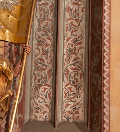 Zdjęcie nr 10: Malowidła z przedstawieniem św. Michała Archanioła pokonującego szatana i św. Jerzego walczącego ze smokiem zostały wykonane na ścianie północnej w dwóch wnękach podokiennych. Obie kompozycje powstały w polach o kształcie wydłużonego prostokąta. Święty Michał Archanioł został przedstawiony frontalnie w całej postaci z głową w ujęciu trzech czwartych w lewo. W uniesionej ponad głowę prawej ręce trzyma włócznię, wbitą w deptanego pod nogami szatana, którą jednocześnie podtrzymuje lewą ręką. Z tyłu widać jego rozpostarte skrzydła, skrzyżowane nad głową. Archanioł ma owalną twarz o wydatnym nosie oraz jasne włosy spływające na ramiona. Ubrany jest w przepasaną  tunikę sięgającą goleni i w narzucony na ramiona płaszcz. Postać diabła pod jego stopami obutymi w buty jest prawie niewidoczna. W podobny sposób przedstawiono św. Jerzego. Święty został ukazany frontalnie na rozstawionych nogach w całej postaci ujętej w kontrapoście z głową w trzech czwartych skierowaną w lewo. Prawą rękę ma uniesioną ponad głową i zgiętą w łokciu. Trzyma w niej lancę podtrzymywaną lewą ręką. Pod jego stopami widoczny jest zarys zwiniętego smoka o zarysie bazyliszka. Św. Jerzy ubrany jest w strój rycerski. Na głowie ma spiczasto zakończony hełm z kolczugą. Ciało okrywa mocno taliowana w pasie tunika zdobiona u dołu ornamentem z motywem trójkąta równoramiennego. Na nogach ma założone wysokie buty. W obu przedstawieniach widoczne są ciemne ugry, ciemna czerwień, żółcień, błękit i brąz. Tła są szare. 