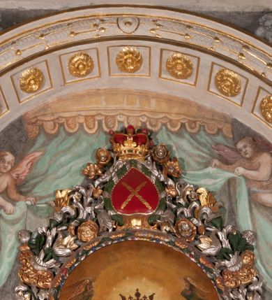 Zdjęcie nr 32: Obraz w formie stojącego prostokąta zamkniętego łukiem półkolistym. W centrum siedząca Matka Boska ukazana do kolan, frontalnie, lewym ramieniem obejmuje siedzące na jej kolanie Dzieciątko, w prawej ręce z przodu trzyma berło. Twarz ma owalną, o delikatnych rysach, dużych, migdałowatych oczach, długim, prostym nosie i niewielkich, pełnych ustach, okoloną długimi brązowymi włosami, opadającymi na ramiona. Ubrana jest w różową suknię, marszczoną przy dekolcie, z dekoracyjną lamówką oraz w obfity ciemnoniebieski płaszcz zarzucony na ramiona i opadający na kolana, ze złoconą lamówką i gwiazdą na prawym barku. Na głowie ma koronę otwartą, wokół nimb gwieździsty. Dzieciątko w pozycji siedzącej, zwrócone w trzech czwartych w prawo, w lewej ręce trzyma jabłko królewskie, prawą unosi w geście błogosławieństwa. Twarz ma kwadratową o pełnych policzkach, migdałowatych oczach, małym nosie i pełnych ustach, okoloną jasnymi, kręconymi włosami. Ubrane jest w fioletową tunikę oraz czerwony płaszcz przerzucony przez kolana. Na głowie ma koronę otwartą. Postacie ukazane na pomarańczowo-brązowym tle, koncentrycznie rozjaśnionym wokół głowy Marii, którą flankują dwa niewielkie anioły z rękami złożonymi w geście modlitwy. Obraz ujęty ornamentalną ramą utworzoną z wici roślinnej przeplatanej kwiatami, tworzącą nad łukiem szczyt z medalionem w wieńcu laurowym, ze skrzyżowanymi laskami marszałkowskimi w polu zwieńczonym koroną zamkniętą. W dolnej partii ramy płaskorzeźbione przedstawienie Chrystusa i Samarytanki przy studni z inicjałami „T(homas) O(linski) D(ecanus) W(ielicensis) P(lebanus) N(iepolomicensis) V(ir) I(llustris)”. 

