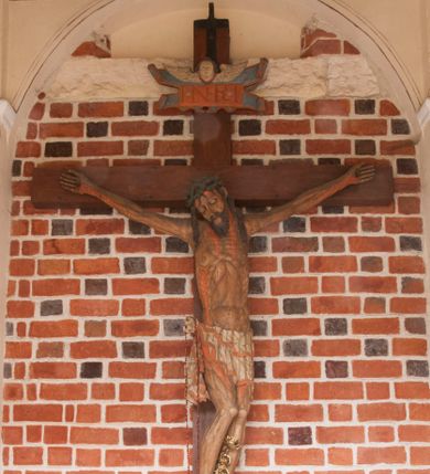 Zdjęcie nr 3: Pełnoplastyczna figura Chrystusa umarłego przybitego do krzyża o prostym zakończeniu ramion trzema gwoździami. Postać w lekkim zwisie, o szeroko rozłożonych ramionach, głowa opada na prawe ramię, nogi ugięte w kolanach, stopy skrzyżowane w układzie prawa na lewą. Jezus ma pociągłą twarz o dużych, zamkniętych oczach, prostym nosie i rozchylonych, wąskich ustach, okoloną falowaną, rozdwajającą się brodą oraz falowanymi, długimi włosami w formie pojedynczych pukli. Na głowie szeroka korona cierniowa. Ciało smukłe, wydłużone, z podkreśloną muskulaturą klatki piersiowej, pokryte siecią żył, z brodzącą rzeźbionymi kroplami krwi raną w boku. Perizonium sięgające połowy uda, zawiązane na prawym biodrze ze zwisem tkaniny, lamowane frędzlą. Ciało o ciemnej karnacji, z zaznaczonymi śladami męki, perizonium polichromowane na biało z wzorem w pasy. Pionowa belka krzyża zakończona rollwerkiem z uskrzydloną główką anielską z napisem „INRI”. 
Krucyfiks umieszczony w arkadzie ujętej pilastrami, dźwigającymi belkowanie. W partii fryzu napis „AGNUS MANSUETUS PORTATUR AD VICTIMAN”.
