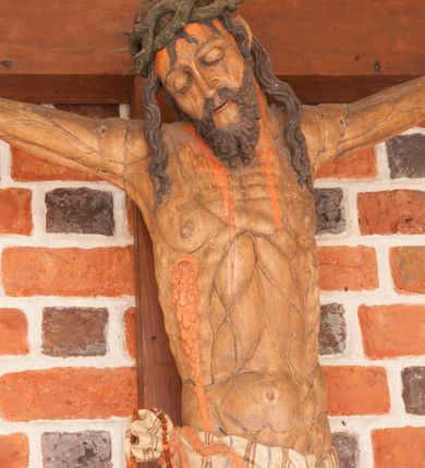 Zdjęcie nr 5: Pełnoplastyczna figura Chrystusa umarłego przybitego do krzyża o prostym zakończeniu ramion trzema gwoździami. Postać w lekkim zwisie, o szeroko rozłożonych ramionach, głowa opada na prawe ramię, nogi ugięte w kolanach, stopy skrzyżowane w układzie prawa na lewą. Jezus ma pociągłą twarz o dużych, zamkniętych oczach, prostym nosie i rozchylonych, wąskich ustach, okoloną falowaną, rozdwajającą się brodą oraz falowanymi, długimi włosami w formie pojedynczych pukli. Na głowie szeroka korona cierniowa. Ciało smukłe, wydłużone, z podkreśloną muskulaturą klatki piersiowej, pokryte siecią żył, z brodzącą rzeźbionymi kroplami krwi raną w boku. Perizonium sięgające połowy uda, zawiązane na prawym biodrze ze zwisem tkaniny, lamowane frędzlą. Ciało o ciemnej karnacji, z zaznaczonymi śladami męki, perizonium polichromowane na biało z wzorem w pasy. Pionowa belka krzyża zakończona rollwerkiem z uskrzydloną główką anielską z napisem „INRI”. 
Krucyfiks umieszczony w arkadzie ujętej pilastrami, dźwigającymi belkowanie. W partii fryzu napis „AGNUS MANSUETUS PORTATUR AD VICTIMAN”.