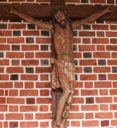 Zdjęcie nr 2: Pełnoplastyczna figura Chrystusa umarłego przybitego do krzyża o prostym zakończeniu ramion trzema gwoździami. Postać w lekkim zwisie, o szeroko rozłożonych ramionach, głowa opada na prawe ramię, nogi ugięte w kolanach, stopy skrzyżowane w układzie prawa na lewą. Jezus ma pociągłą twarz o dużych, zamkniętych oczach, prostym nosie i rozchylonych, wąskich ustach, okoloną falowaną, rozdwajającą się brodą oraz falowanymi, długimi włosami w formie pojedynczych pukli. Na głowie szeroka korona cierniowa. Ciało smukłe, wydłużone, z podkreśloną muskulaturą klatki piersiowej, pokryte siecią żył, z brodzącą rzeźbionymi kroplami krwi raną w boku. Perizonium sięgające połowy uda, zawiązane na prawym biodrze ze zwisem tkaniny, lamowane frędzlą. Ciało o ciemnej karnacji, z zaznaczonymi śladami męki, perizonium polichromowane na biało z wzorem w pasy. Pionowa belka krzyża zakończona rollwerkiem z uskrzydloną główką anielską z napisem „INRI”. 
Krucyfiks umieszczony w arkadzie ujętej pilastrami, dźwigającymi belkowanie. W partii fryzu napis „AGNUS MANSUETUS PORTATUR AD VICTIMAN”.