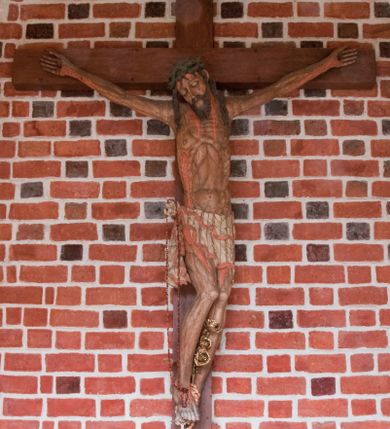 Zdjęcie nr 1: Pełnoplastyczna figura Chrystusa umarłego przybitego do krzyża o prostym zakończeniu ramion trzema gwoździami. Postać w lekkim zwisie, o szeroko rozłożonych ramionach, głowa opada na prawe ramię, nogi ugięte w kolanach, stopy skrzyżowane w układzie prawa na lewą. Jezus ma pociągłą twarz o dużych, zamkniętych oczach, prostym nosie i rozchylonych, wąskich ustach, okoloną falowaną, rozdwajającą się brodą oraz falowanymi, długimi włosami w formie pojedynczych pukli. Na głowie szeroka korona cierniowa. Ciało smukłe, wydłużone, z podkreśloną muskulaturą klatki piersiowej, pokryte siecią żył, z brodzącą rzeźbionymi kroplami krwi raną w boku. Perizonium sięgające połowy uda, zawiązane na prawym biodrze ze zwisem tkaniny, lamowane frędzlą. Ciało o ciemnej karnacji, z zaznaczonymi śladami męki, perizonium polichromowane na biało z wzorem w pasy. Pionowa belka krzyża zakończona rollwerkiem z uskrzydloną główką anielską z napisem „INRI”. 
Krucyfiks umieszczony w arkadzie ujętej pilastrami, dźwigającymi belkowanie. W partii fryzu napis „AGNUS MANSUETUS PORTATUR AD VICTIMAN”.