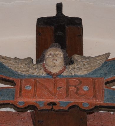 Zdjęcie nr 6: Pełnoplastyczna figura Chrystusa umarłego przybitego do krzyża o prostym zakończeniu ramion trzema gwoździami. Postać w lekkim zwisie, o szeroko rozłożonych ramionach, głowa opada na prawe ramię, nogi ugięte w kolanach, stopy skrzyżowane w układzie prawa na lewą. Jezus ma pociągłą twarz o dużych, zamkniętych oczach, prostym nosie i rozchylonych, wąskich ustach, okoloną falowaną, rozdwajającą się brodą oraz falowanymi, długimi włosami w formie pojedynczych pukli. Na głowie szeroka korona cierniowa. Ciało smukłe, wydłużone, z podkreśloną muskulaturą klatki piersiowej, pokryte siecią żył, z brodzącą rzeźbionymi kroplami krwi raną w boku. Perizonium sięgające połowy uda, zawiązane na prawym biodrze ze zwisem tkaniny, lamowane frędzlą. Ciało o ciemnej karnacji, z zaznaczonymi śladami męki, perizonium polichromowane na biało z wzorem w pasy. Pionowa belka krzyża zakończona rollwerkiem z uskrzydloną główką anielską z napisem „INRI”. 
Krucyfiks umieszczony w arkadzie ujętej pilastrami, dźwigającymi belkowanie. W partii fryzu napis „AGNUS MANSUETUS PORTATUR AD VICTIMAN”.