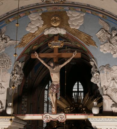 Zdjęcie nr 3: Pełnoplastyczna figura Chrystusa Ukrzyżowanego umieszczona jest na belce tęczowej. Chrystus został ukazany na krzyżu w dużym zwisie na wyprężonych ramionach, tworzących kształt litery „V”. Jego korpus jest wydłużony i esowato wygięty z silnym zaznaczeniem modelunku anatomicznego. Nogi zostały lekko zgięte w kolanach i nieznacznie odchylone w bok, zaś stopy skrzyżowane i przybite jednym gwoździem do krzyża. Wyeksponowano muskulaturę kończyn, żebra i ranę w boku. Głowa Zbawiciela opada na prawe ramię. Jego twarz jest podłużna, z długim, prostym nosem, o zamkniętych oczach i otwartych ustach w grymasie bólu. Otacza ją gęsta, kręcona broda oraz długie, ciemne włosy opadające lokami na oba ramiona. Na głowie widoczna jest cierniowa korona o długich kolcach. Biodra Zbawiciela zostały przepasane długim perizonium sięgającym kolan. Jest ono na środku drapowane za pomocą misowych fałd układających się poprzecznie, zaś po bokach zostało wywinięte za pomocą dwóch festonów opadających kaskadowo wzdłuż ud. Ponad głową Chrystusa umieszczony jest złocony titulus o kształtach barokowych w formie kartusza z napisem „INRI”.Rzeźba jest polichromowana, a perizonium srebrzone z zastosowaniem laserunków.