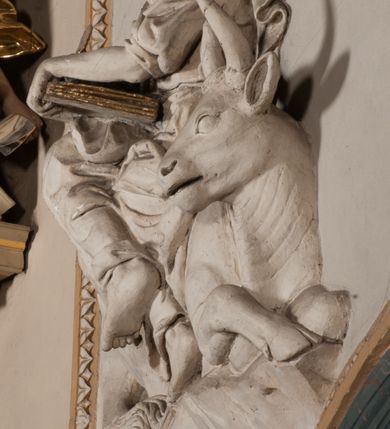 Zdjęcie nr 19: Pełnoplastyczna figura Chrystusa Ukrzyżowanego umieszczona jest na belce tęczowej. Chrystus został ukazany na krzyżu w dużym zwisie na wyprężonych ramionach, tworzących kształt litery „V”. Jego korpus jest wydłużony i esowato wygięty z silnym zaznaczeniem modelunku anatomicznego. Nogi zostały lekko zgięte w kolanach i nieznacznie odchylone w bok, zaś stopy skrzyżowane i przybite jednym gwoździem do krzyża. Wyeksponowano muskulaturę kończyn, żebra i ranę w boku. Głowa Zbawiciela opada na prawe ramię. Jego twarz jest podłużna, z długim, prostym nosem, o zamkniętych oczach i otwartych ustach w grymasie bólu. Otacza ją gęsta, kręcona broda oraz długie, ciemne włosy opadające lokami na oba ramiona. Na głowie widoczna jest cierniowa korona o długich kolcach. Biodra Zbawiciela zostały przepasane długim perizonium sięgającym kolan. Jest ono na środku drapowane za pomocą misowych fałd układających się poprzecznie, zaś po bokach zostało wywinięte za pomocą dwóch festonów opadających kaskadowo wzdłuż ud. Ponad głową Chrystusa umieszczony jest złocony titulus o kształtach barokowych w formie kartusza z napisem „INRI”.Rzeźba jest polichromowana, a perizonium srebrzone z zastosowaniem laserunków.