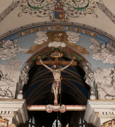 Zdjęcie nr 5: Pełnoplastyczna figura Chrystusa Ukrzyżowanego umieszczona jest na belce tęczowej. Chrystus został ukazany na krzyżu w dużym zwisie na wyprężonych ramionach, tworzących kształt litery „V”. Jego korpus jest wydłużony i esowato wygięty z silnym zaznaczeniem modelunku anatomicznego. Nogi zostały lekko zgięte w kolanach i nieznacznie odchylone w bok, zaś stopy skrzyżowane i przybite jednym gwoździem do krzyża. Wyeksponowano muskulaturę kończyn, żebra i ranę w boku. Głowa Zbawiciela opada na prawe ramię. Jego twarz jest podłużna, z długim, prostym nosem, o zamkniętych oczach i otwartych ustach w grymasie bólu. Otacza ją gęsta, kręcona broda oraz długie, ciemne włosy opadające lokami na oba ramiona. Na głowie widoczna jest cierniowa korona o długich kolcach. Biodra Zbawiciela zostały przepasane długim perizonium sięgającym kolan. Jest ono na środku drapowane za pomocą misowych fałd układających się poprzecznie, zaś po bokach zostało wywinięte za pomocą dwóch festonów opadających kaskadowo wzdłuż ud. Ponad głową Chrystusa umieszczony jest złocony titulus o kształtach barokowych w formie kartusza z napisem „INRI”.Rzeźba jest polichromowana, a perizonium srebrzone z zastosowaniem laserunków.
