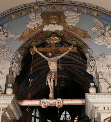 Zdjęcie nr 2: Pełnoplastyczna figura Chrystusa Ukrzyżowanego umieszczona jest na belce tęczowej. Chrystus został ukazany na krzyżu w dużym zwisie na wyprężonych ramionach, tworzących kształt litery „V”. Jego korpus jest wydłużony i esowato wygięty z silnym zaznaczeniem modelunku anatomicznego. Nogi zostały lekko zgięte w kolanach i nieznacznie odchylone w bok, zaś stopy skrzyżowane i przybite jednym gwoździem do krzyża. Wyeksponowano muskulaturę kończyn, żebra i ranę w boku. Głowa Zbawiciela opada na prawe ramię. Jego twarz jest podłużna, z długim, prostym nosem, o zamkniętych oczach i otwartych ustach w grymasie bólu. Otacza ją gęsta, kręcona broda oraz długie, ciemne włosy opadające lokami na oba ramiona. Na głowie widoczna jest cierniowa korona o długich kolcach. Biodra Zbawiciela zostały przepasane długim perizonium sięgającym kolan. Jest ono na środku drapowane za pomocą misowych fałd układających się poprzecznie, zaś po bokach zostało wywinięte za pomocą dwóch festonów opadających kaskadowo wzdłuż ud. Ponad głową Chrystusa umieszczony jest złocony titulus o kształtach barokowych w formie kartusza z napisem „INRI”.Rzeźba jest polichromowana, a perizonium srebrzone z zastosowaniem laserunków.