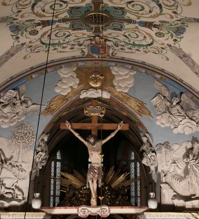 Zdjęcie nr 4: Pełnoplastyczna figura Chrystusa Ukrzyżowanego umieszczona jest na belce tęczowej. Chrystus został ukazany na krzyżu w dużym zwisie na wyprężonych ramionach, tworzących kształt litery „V”. Jego korpus jest wydłużony i esowato wygięty z silnym zaznaczeniem modelunku anatomicznego. Nogi zostały lekko zgięte w kolanach i nieznacznie odchylone w bok, zaś stopy skrzyżowane i przybite jednym gwoździem do krzyża. Wyeksponowano muskulaturę kończyn, żebra i ranę w boku. Głowa Zbawiciela opada na prawe ramię. Jego twarz jest podłużna, z długim, prostym nosem, o zamkniętych oczach i otwartych ustach w grymasie bólu. Otacza ją gęsta, kręcona broda oraz długie, ciemne włosy opadające lokami na oba ramiona. Na głowie widoczna jest cierniowa korona o długich kolcach. Biodra Zbawiciela zostały przepasane długim perizonium sięgającym kolan. Jest ono na środku drapowane za pomocą misowych fałd układających się poprzecznie, zaś po bokach zostało wywinięte za pomocą dwóch festonów opadających kaskadowo wzdłuż ud. Ponad głową Chrystusa umieszczony jest złocony titulus o kształtach barokowych w formie kartusza z napisem „INRI”.Rzeźba jest polichromowana, a perizonium srebrzone z zastosowaniem laserunków.