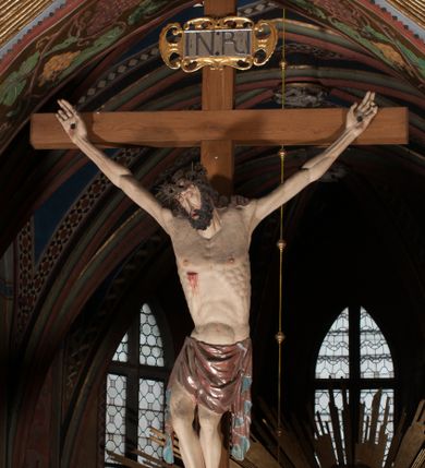 Zdjęcie nr 1: Pełnoplastyczna figura Chrystusa Ukrzyżowanego umieszczona jest na belce tęczowej. Chrystus został ukazany na krzyżu w dużym zwisie na wyprężonych ramionach, tworzących kształt litery „V”. Jego korpus jest wydłużony i esowato wygięty z silnym zaznaczeniem modelunku anatomicznego. Nogi zostały lekko zgięte w kolanach i nieznacznie odchylone w bok, zaś stopy skrzyżowane i przybite jednym gwoździem do krzyża. Wyeksponowano muskulaturę kończyn, żebra i ranę w boku. Głowa Zbawiciela opada na prawe ramię. Jego twarz jest podłużna, z długim, prostym nosem, o zamkniętych oczach i otwartych ustach w grymasie bólu. Otacza ją gęsta, kręcona broda oraz długie, ciemne włosy opadające lokami na oba ramiona. Na głowie widoczna jest cierniowa korona o długich kolcach. Biodra Zbawiciela zostały przepasane długim perizonium sięgającym kolan. Jest ono na środku drapowane za pomocą misowych fałd układających się poprzecznie, zaś po bokach zostało wywinięte za pomocą dwóch festonów opadających kaskadowo wzdłuż ud. Ponad głową Chrystusa umieszczony jest złocony titulus o kształtach barokowych w formie kartusza z napisem „INRI”.Rzeźba jest polichromowana, a perizonium srebrzone z zastosowaniem laserunków.