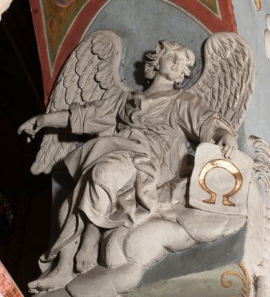 Zdjęcie nr 23: Pełnoplastyczna figura Chrystusa Ukrzyżowanego umieszczona jest na belce tęczowej. Chrystus został ukazany na krzyżu w dużym zwisie na wyprężonych ramionach, tworzących kształt litery „V”. Jego korpus jest wydłużony i esowato wygięty z silnym zaznaczeniem modelunku anatomicznego. Nogi zostały lekko zgięte w kolanach i nieznacznie odchylone w bok, zaś stopy skrzyżowane i przybite jednym gwoździem do krzyża. Wyeksponowano muskulaturę kończyn, żebra i ranę w boku. Głowa Zbawiciela opada na prawe ramię. Jego twarz jest podłużna, z długim, prostym nosem, o zamkniętych oczach i otwartych ustach w grymasie bólu. Otacza ją gęsta, kręcona broda oraz długie, ciemne włosy opadające lokami na oba ramiona. Na głowie widoczna jest cierniowa korona o długich kolcach. Biodra Zbawiciela zostały przepasane długim perizonium sięgającym kolan. Jest ono na środku drapowane za pomocą misowych fałd układających się poprzecznie, zaś po bokach zostało wywinięte za pomocą dwóch festonów opadających kaskadowo wzdłuż ud. Ponad głową Chrystusa umieszczony jest złocony titulus o kształtach barokowych w formie kartusza z napisem „INRI”.Rzeźba jest polichromowana, a perizonium srebrzone z zastosowaniem laserunków.