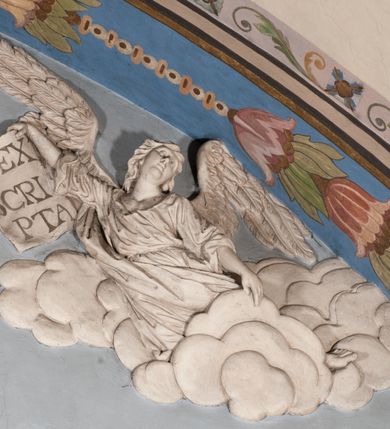 Zdjęcie nr 28: Pełnoplastyczna figura Chrystusa Ukrzyżowanego umieszczona jest na belce tęczowej. Chrystus został ukazany na krzyżu w dużym zwisie na wyprężonych ramionach, tworzących kształt litery „V”. Jego korpus jest wydłużony i esowato wygięty z silnym zaznaczeniem modelunku anatomicznego. Nogi zostały lekko zgięte w kolanach i nieznacznie odchylone w bok, zaś stopy skrzyżowane i przybite jednym gwoździem do krzyża. Wyeksponowano muskulaturę kończyn, żebra i ranę w boku. Głowa Zbawiciela opada na prawe ramię. Jego twarz jest podłużna, z długim, prostym nosem, o zamkniętych oczach i otwartych ustach w grymasie bólu. Otacza ją gęsta, kręcona broda oraz długie, ciemne włosy opadające lokami na oba ramiona. Na głowie widoczna jest cierniowa korona o długich kolcach. Biodra Zbawiciela zostały przepasane długim perizonium sięgającym kolan. Jest ono na środku drapowane za pomocą misowych fałd układających się poprzecznie, zaś po bokach zostało wywinięte za pomocą dwóch festonów opadających kaskadowo wzdłuż ud. Ponad głową Chrystusa umieszczony jest złocony titulus o kształtach barokowych w formie kartusza z napisem „INRI”.Rzeźba jest polichromowana, a perizonium srebrzone z zastosowaniem laserunków.