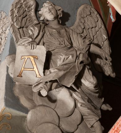 Zdjęcie nr 25: Pełnoplastyczna figura Chrystusa Ukrzyżowanego umieszczona jest na belce tęczowej. Chrystus został ukazany na krzyżu w dużym zwisie na wyprężonych ramionach, tworzących kształt litery „V”. Jego korpus jest wydłużony i esowato wygięty z silnym zaznaczeniem modelunku anatomicznego. Nogi zostały lekko zgięte w kolanach i nieznacznie odchylone w bok, zaś stopy skrzyżowane i przybite jednym gwoździem do krzyża. Wyeksponowano muskulaturę kończyn, żebra i ranę w boku. Głowa Zbawiciela opada na prawe ramię. Jego twarz jest podłużna, z długim, prostym nosem, o zamkniętych oczach i otwartych ustach w grymasie bólu. Otacza ją gęsta, kręcona broda oraz długie, ciemne włosy opadające lokami na oba ramiona. Na głowie widoczna jest cierniowa korona o długich kolcach. Biodra Zbawiciela zostały przepasane długim perizonium sięgającym kolan. Jest ono na środku drapowane za pomocą misowych fałd układających się poprzecznie, zaś po bokach zostało wywinięte za pomocą dwóch festonów opadających kaskadowo wzdłuż ud. Ponad głową Chrystusa umieszczony jest złocony titulus o kształtach barokowych w formie kartusza z napisem „INRI”.Rzeźba jest polichromowana, a perizonium srebrzone z zastosowaniem laserunków.