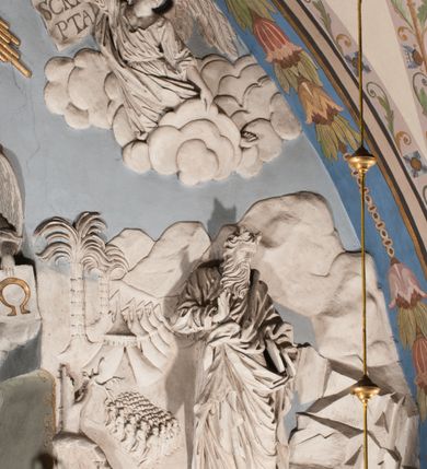 Zdjęcie nr 33: Pełnoplastyczna figura Chrystusa Ukrzyżowanego umieszczona jest na belce tęczowej. Chrystus został ukazany na krzyżu w dużym zwisie na wyprężonych ramionach, tworzących kształt litery „V”. Jego korpus jest wydłużony i esowato wygięty z silnym zaznaczeniem modelunku anatomicznego. Nogi zostały lekko zgięte w kolanach i nieznacznie odchylone w bok, zaś stopy skrzyżowane i przybite jednym gwoździem do krzyża. Wyeksponowano muskulaturę kończyn, żebra i ranę w boku. Głowa Zbawiciela opada na prawe ramię. Jego twarz jest podłużna, z długim, prostym nosem, o zamkniętych oczach i otwartych ustach w grymasie bólu. Otacza ją gęsta, kręcona broda oraz długie, ciemne włosy opadające lokami na oba ramiona. Na głowie widoczna jest cierniowa korona o długich kolcach. Biodra Zbawiciela zostały przepasane długim perizonium sięgającym kolan. Jest ono na środku drapowane za pomocą misowych fałd układających się poprzecznie, zaś po bokach zostało wywinięte za pomocą dwóch festonów opadających kaskadowo wzdłuż ud. Ponad głową Chrystusa umieszczony jest złocony titulus o kształtach barokowych w formie kartusza z napisem „INRI”.Rzeźba jest polichromowana, a perizonium srebrzone z zastosowaniem laserunków.