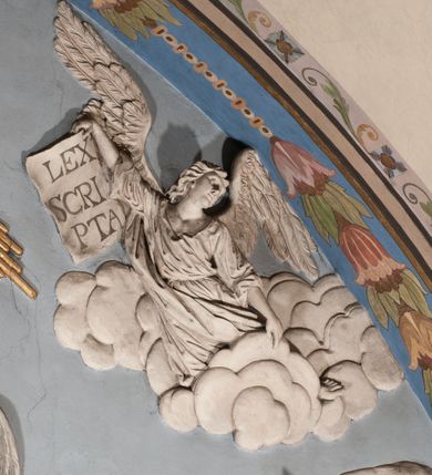 Zdjęcie nr 50: Pełnoplastyczna figura Chrystusa Ukrzyżowanego umieszczona jest na belce tęczowej. Chrystus został ukazany na krzyżu w dużym zwisie na wyprężonych ramionach, tworzących kształt litery „V”. Jego korpus jest wydłużony i esowato wygięty z silnym zaznaczeniem modelunku anatomicznego. Nogi zostały lekko zgięte w kolanach i nieznacznie odchylone w bok, zaś stopy skrzyżowane i przybite jednym gwoździem do krzyża. Wyeksponowano muskulaturę kończyn, żebra i ranę w boku. Głowa Zbawiciela opada na prawe ramię. Jego twarz jest podłużna, z długim, prostym nosem, o zamkniętych oczach i otwartych ustach w grymasie bólu. Otacza ją gęsta, kręcona broda oraz długie, ciemne włosy opadające lokami na oba ramiona. Na głowie widoczna jest cierniowa korona o długich kolcach. Biodra Zbawiciela zostały przepasane długim perizonium sięgającym kolan. Jest ono na środku drapowane za pomocą misowych fałd układających się poprzecznie, zaś po bokach zostało wywinięte za pomocą dwóch festonów opadających kaskadowo wzdłuż ud. Ponad głową Chrystusa umieszczony jest złocony titulus o kształtach barokowych w formie kartusza z napisem „INRI”.Rzeźba jest polichromowana, a perizonium srebrzone z zastosowaniem laserunków.