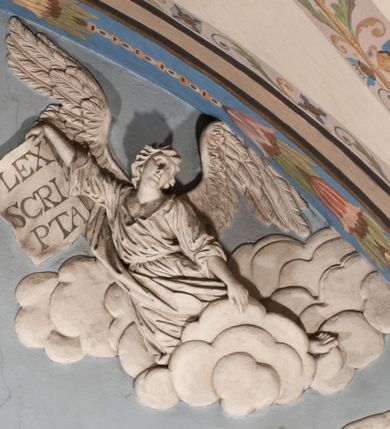 Zdjęcie nr 51: Pełnoplastyczna figura Chrystusa Ukrzyżowanego umieszczona jest na belce tęczowej. Chrystus został ukazany na krzyżu w dużym zwisie na wyprężonych ramionach, tworzących kształt litery „V”. Jego korpus jest wydłużony i esowato wygięty z silnym zaznaczeniem modelunku anatomicznego. Nogi zostały lekko zgięte w kolanach i nieznacznie odchylone w bok, zaś stopy skrzyżowane i przybite jednym gwoździem do krzyża. Wyeksponowano muskulaturę kończyn, żebra i ranę w boku. Głowa Zbawiciela opada na prawe ramię. Jego twarz jest podłużna, z długim, prostym nosem, o zamkniętych oczach i otwartych ustach w grymasie bólu. Otacza ją gęsta, kręcona broda oraz długie, ciemne włosy opadające lokami na oba ramiona. Na głowie widoczna jest cierniowa korona o długich kolcach. Biodra Zbawiciela zostały przepasane długim perizonium sięgającym kolan. Jest ono na środku drapowane za pomocą misowych fałd układających się poprzecznie, zaś po bokach zostało wywinięte za pomocą dwóch festonów opadających kaskadowo wzdłuż ud. Ponad głową Chrystusa umieszczony jest złocony titulus o kształtach barokowych w formie kartusza z napisem „INRI”.Rzeźba jest polichromowana, a perizonium srebrzone z zastosowaniem laserunków.