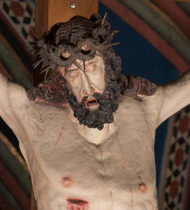 Zdjęcie nr 10: Pełnoplastyczna figura Chrystusa Ukrzyżowanego umieszczona jest na belce tęczowej. Chrystus został ukazany na krzyżu w dużym zwisie na wyprężonych ramionach, tworzących kształt litery „V”. Jego korpus jest wydłużony i esowato wygięty z silnym zaznaczeniem modelunku anatomicznego. Nogi zostały lekko zgięte w kolanach i nieznacznie odchylone w bok, zaś stopy skrzyżowane i przybite jednym gwoździem do krzyża. Wyeksponowano muskulaturę kończyn, żebra i ranę w boku. Głowa Zbawiciela opada na prawe ramię. Jego twarz jest podłużna, z długim, prostym nosem, o zamkniętych oczach i otwartych ustach w grymasie bólu. Otacza ją gęsta, kręcona broda oraz długie, ciemne włosy opadające lokami na oba ramiona. Na głowie widoczna jest cierniowa korona o długich kolcach. Biodra Zbawiciela zostały przepasane długim perizonium sięgającym kolan. Jest ono na środku drapowane za pomocą misowych fałd układających się poprzecznie, zaś po bokach zostało wywinięte za pomocą dwóch festonów opadających kaskadowo wzdłuż ud. Ponad głową Chrystusa umieszczony jest złocony titulus o kształtach barokowych w formie kartusza z napisem „INRI”.Rzeźba jest polichromowana, a perizonium srebrzone z zastosowaniem laserunków.