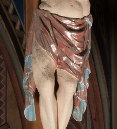 Zdjęcie nr 11: Pełnoplastyczna figura Chrystusa Ukrzyżowanego umieszczona jest na belce tęczowej. Chrystus został ukazany na krzyżu w dużym zwisie na wyprężonych ramionach, tworzących kształt litery „V”. Jego korpus jest wydłużony i esowato wygięty z silnym zaznaczeniem modelunku anatomicznego. Nogi zostały lekko zgięte w kolanach i nieznacznie odchylone w bok, zaś stopy skrzyżowane i przybite jednym gwoździem do krzyża. Wyeksponowano muskulaturę kończyn, żebra i ranę w boku. Głowa Zbawiciela opada na prawe ramię. Jego twarz jest podłużna, z długim, prostym nosem, o zamkniętych oczach i otwartych ustach w grymasie bólu. Otacza ją gęsta, kręcona broda oraz długie, ciemne włosy opadające lokami na oba ramiona. Na głowie widoczna jest cierniowa korona o długich kolcach. Biodra Zbawiciela zostały przepasane długim perizonium sięgającym kolan. Jest ono na środku drapowane za pomocą misowych fałd układających się poprzecznie, zaś po bokach zostało wywinięte za pomocą dwóch festonów opadających kaskadowo wzdłuż ud. Ponad głową Chrystusa umieszczony jest złocony titulus o kształtach barokowych w formie kartusza z napisem „INRI”.Rzeźba jest polichromowana, a perizonium srebrzone z zastosowaniem laserunków.