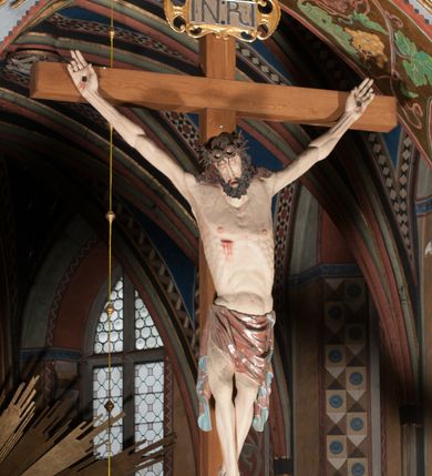 Zdjęcie nr 6: Pełnoplastyczna figura Chrystusa Ukrzyżowanego umieszczona jest na belce tęczowej. Chrystus został ukazany na krzyżu w dużym zwisie na wyprężonych ramionach, tworzących kształt litery „V”. Jego korpus jest wydłużony i esowato wygięty z silnym zaznaczeniem modelunku anatomicznego. Nogi zostały lekko zgięte w kolanach i nieznacznie odchylone w bok, zaś stopy skrzyżowane i przybite jednym gwoździem do krzyża. Wyeksponowano muskulaturę kończyn, żebra i ranę w boku. Głowa Zbawiciela opada na prawe ramię. Jego twarz jest podłużna, z długim, prostym nosem, o zamkniętych oczach i otwartych ustach w grymasie bólu. Otacza ją gęsta, kręcona broda oraz długie, ciemne włosy opadające lokami na oba ramiona. Na głowie widoczna jest cierniowa korona o długich kolcach. Biodra Zbawiciela zostały przepasane długim perizonium sięgającym kolan. Jest ono na środku drapowane za pomocą misowych fałd układających się poprzecznie, zaś po bokach zostało wywinięte za pomocą dwóch festonów opadających kaskadowo wzdłuż ud. Ponad głową Chrystusa umieszczony jest złocony titulus o kształtach barokowych w formie kartusza z napisem „INRI”.Rzeźba jest polichromowana, a perizonium srebrzone z zastosowaniem laserunków.