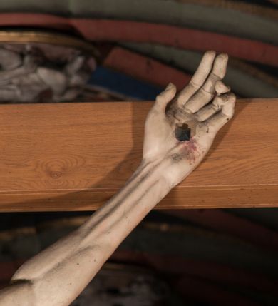 Zdjęcie nr 15: Pełnoplastyczna figura Chrystusa Ukrzyżowanego umieszczona jest na belce tęczowej. Chrystus został ukazany na krzyżu w dużym zwisie na wyprężonych ramionach, tworzących kształt litery „V”. Jego korpus jest wydłużony i esowato wygięty z silnym zaznaczeniem modelunku anatomicznego. Nogi zostały lekko zgięte w kolanach i nieznacznie odchylone w bok, zaś stopy skrzyżowane i przybite jednym gwoździem do krzyża. Wyeksponowano muskulaturę kończyn, żebra i ranę w boku. Głowa Zbawiciela opada na prawe ramię. Jego twarz jest podłużna, z długim, prostym nosem, o zamkniętych oczach i otwartych ustach w grymasie bólu. Otacza ją gęsta, kręcona broda oraz długie, ciemne włosy opadające lokami na oba ramiona. Na głowie widoczna jest cierniowa korona o długich kolcach. Biodra Zbawiciela zostały przepasane długim perizonium sięgającym kolan. Jest ono na środku drapowane za pomocą misowych fałd układających się poprzecznie, zaś po bokach zostało wywinięte za pomocą dwóch festonów opadających kaskadowo wzdłuż ud. Ponad głową Chrystusa umieszczony jest złocony titulus o kształtach barokowych w formie kartusza z napisem „INRI”.Rzeźba jest polichromowana, a perizonium srebrzone z zastosowaniem laserunków.