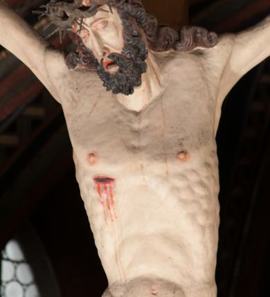 Zdjęcie nr 7: Pełnoplastyczna figura Chrystusa Ukrzyżowanego umieszczona jest na belce tęczowej. Chrystus został ukazany na krzyżu w dużym zwisie na wyprężonych ramionach, tworzących kształt litery „V”. Jego korpus jest wydłużony i esowato wygięty z silnym zaznaczeniem modelunku anatomicznego. Nogi zostały lekko zgięte w kolanach i nieznacznie odchylone w bok, zaś stopy skrzyżowane i przybite jednym gwoździem do krzyża. Wyeksponowano muskulaturę kończyn, żebra i ranę w boku. Głowa Zbawiciela opada na prawe ramię. Jego twarz jest podłużna, z długim, prostym nosem, o zamkniętych oczach i otwartych ustach w grymasie bólu. Otacza ją gęsta, kręcona broda oraz długie, ciemne włosy opadające lokami na oba ramiona. Na głowie widoczna jest cierniowa korona o długich kolcach. Biodra Zbawiciela zostały przepasane długim perizonium sięgającym kolan. Jest ono na środku drapowane za pomocą misowych fałd układających się poprzecznie, zaś po bokach zostało wywinięte za pomocą dwóch festonów opadających kaskadowo wzdłuż ud. Ponad głową Chrystusa umieszczony jest złocony titulus o kształtach barokowych w formie kartusza z napisem „INRI”.Rzeźba jest polichromowana, a perizonium srebrzone z zastosowaniem laserunków.