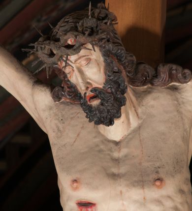 Zdjęcie nr 9: Pełnoplastyczna figura Chrystusa Ukrzyżowanego umieszczona jest na belce tęczowej. Chrystus został ukazany na krzyżu w dużym zwisie na wyprężonych ramionach, tworzących kształt litery „V”. Jego korpus jest wydłużony i esowato wygięty z silnym zaznaczeniem modelunku anatomicznego. Nogi zostały lekko zgięte w kolanach i nieznacznie odchylone w bok, zaś stopy skrzyżowane i przybite jednym gwoździem do krzyża. Wyeksponowano muskulaturę kończyn, żebra i ranę w boku. Głowa Zbawiciela opada na prawe ramię. Jego twarz jest podłużna, z długim, prostym nosem, o zamkniętych oczach i otwartych ustach w grymasie bólu. Otacza ją gęsta, kręcona broda oraz długie, ciemne włosy opadające lokami na oba ramiona. Na głowie widoczna jest cierniowa korona o długich kolcach. Biodra Zbawiciela zostały przepasane długim perizonium sięgającym kolan. Jest ono na środku drapowane za pomocą misowych fałd układających się poprzecznie, zaś po bokach zostało wywinięte za pomocą dwóch festonów opadających kaskadowo wzdłuż ud. Ponad głową Chrystusa umieszczony jest złocony titulus o kształtach barokowych w formie kartusza z napisem „INRI”.Rzeźba jest polichromowana, a perizonium srebrzone z zastosowaniem laserunków.