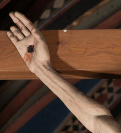 Zdjęcie nr 14: Pełnoplastyczna figura Chrystusa Ukrzyżowanego umieszczona jest na belce tęczowej. Chrystus został ukazany na krzyżu w dużym zwisie na wyprężonych ramionach, tworzących kształt litery „V”. Jego korpus jest wydłużony i esowato wygięty z silnym zaznaczeniem modelunku anatomicznego. Nogi zostały lekko zgięte w kolanach i nieznacznie odchylone w bok, zaś stopy skrzyżowane i przybite jednym gwoździem do krzyża. Wyeksponowano muskulaturę kończyn, żebra i ranę w boku. Głowa Zbawiciela opada na prawe ramię. Jego twarz jest podłużna, z długim, prostym nosem, o zamkniętych oczach i otwartych ustach w grymasie bólu. Otacza ją gęsta, kręcona broda oraz długie, ciemne włosy opadające lokami na oba ramiona. Na głowie widoczna jest cierniowa korona o długich kolcach. Biodra Zbawiciela zostały przepasane długim perizonium sięgającym kolan. Jest ono na środku drapowane za pomocą misowych fałd układających się poprzecznie, zaś po bokach zostało wywinięte za pomocą dwóch festonów opadających kaskadowo wzdłuż ud. Ponad głową Chrystusa umieszczony jest złocony titulus o kształtach barokowych w formie kartusza z napisem „INRI”.Rzeźba jest polichromowana, a perizonium srebrzone z zastosowaniem laserunków.