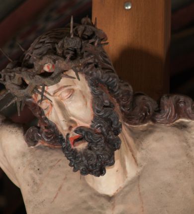 Zdjęcie nr 8: Pełnoplastyczna figura Chrystusa Ukrzyżowanego umieszczona jest na belce tęczowej. Chrystus został ukazany na krzyżu w dużym zwisie na wyprężonych ramionach, tworzących kształt litery „V”. Jego korpus jest wydłużony i esowato wygięty z silnym zaznaczeniem modelunku anatomicznego. Nogi zostały lekko zgięte w kolanach i nieznacznie odchylone w bok, zaś stopy skrzyżowane i przybite jednym gwoździem do krzyża. Wyeksponowano muskulaturę kończyn, żebra i ranę w boku. Głowa Zbawiciela opada na prawe ramię. Jego twarz jest podłużna, z długim, prostym nosem, o zamkniętych oczach i otwartych ustach w grymasie bólu. Otacza ją gęsta, kręcona broda oraz długie, ciemne włosy opadające lokami na oba ramiona. Na głowie widoczna jest cierniowa korona o długich kolcach. Biodra Zbawiciela zostały przepasane długim perizonium sięgającym kolan. Jest ono na środku drapowane za pomocą misowych fałd układających się poprzecznie, zaś po bokach zostało wywinięte za pomocą dwóch festonów opadających kaskadowo wzdłuż ud. Ponad głową Chrystusa umieszczony jest złocony titulus o kształtach barokowych w formie kartusza z napisem „INRI”.Rzeźba jest polichromowana, a perizonium srebrzone z zastosowaniem laserunków.