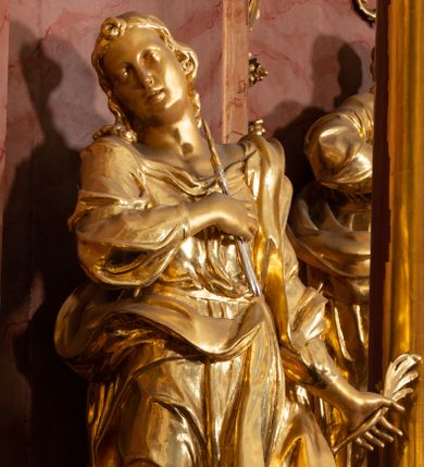 Zdjęcie nr 1: Rzeźba drążona, ustawiona na podstawie na planie kwadratu. Święta ukazana frontalnie, w kontrapoście, o esowato wygiętej sylwetce, z głową przechyloną na prawe ramię. Prawa ręka na piersi, w dłoni trzyma obcęgi zwieńczone lilią heraldyczną, lewa odchylona od boku, w dłoni ma liść palmy. Twarz ma owalną, o wydatnym podbródku, migdałowatych oczach, o głęboko rzeźbionych kącikach, prostym nosie i pełnych, rozchylonych ustach. Włosy ma długie, kręcone, sięgające ramion, zawinięte nad czołem. Ubrana jest w suknię przewiązaną w talii, z dekoltem odsłaniającym ramiona, ściśle przylegającą do ciała oraz w płaszcz zarzucony na lewe ramię i zawiązany z przodu postaci. Figura w całości złocona.