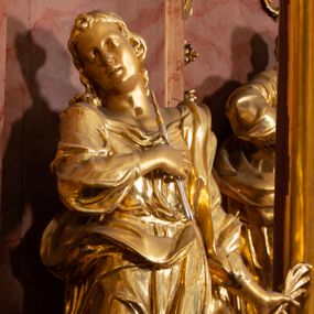 Zdjęcie nr 1: Rzeźba drążona, ustawiona na podstawie na planie kwadratu. Święta ukazana frontalnie, w kontrapoście, o esowato wygiętej sylwetce, z głową przechyloną na prawe ramię. Prawa ręka na piersi, w dłoni trzyma obcęgi zwieńczone lilią heraldyczną, lewa odchylona od boku, w dłoni ma liść palmy. Twarz ma owalną, o wydatnym podbródku, migdałowatych oczach, o głęboko rzeźbionych kącikach, prostym nosie i pełnych, rozchylonych ustach. Włosy ma długie, kręcone, sięgające ramion, zawinięte nad czołem. Ubrana jest w suknię przewiązaną w talii, z dekoltem odsłaniającym ramiona, ściśle przylegającą do ciała oraz w płaszcz zarzucony na lewe ramię i zawiązany z przodu postaci. Figura w całości złocona.