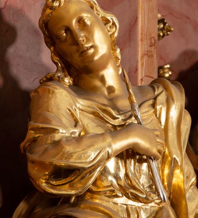 Zdjęcie nr 7: Rzeźba drążona, ustawiona na podstawie na planie kwadratu. Święta ukazana frontalnie, w kontrapoście, o esowato wygiętej sylwetce, z głową przechyloną na prawe ramię. Prawa ręka na piersi, w dłoni trzyma obcęgi zwieńczone lilią heraldyczną, lewa odchylona od boku, w dłoni ma liść palmy. Twarz ma owalną, o wydatnym podbródku, migdałowatych oczach, o głęboko rzeźbionych kącikach, prostym nosie i pełnych, rozchylonych ustach. Włosy ma długie, kręcone, sięgające ramion, zawinięte nad czołem. Ubrana jest w suknię przewiązaną w talii, z dekoltem odsłaniającym ramiona, ściśle przylegającą do ciała oraz w płaszcz zarzucony na lewe ramię i zawiązany z przodu postaci. Figura w całości złocona.