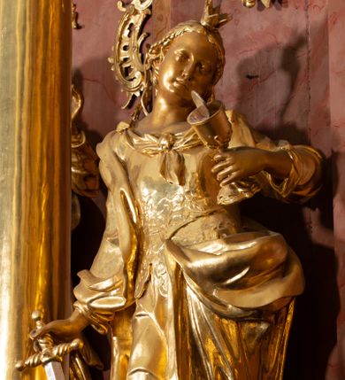 Zdjęcie nr 1: Rzeźba drążona, ustawiona na podstawie. Święta ukazana frontalnie, w kontrapoście, o esowato wygiętej sylwetce, z silnie wypchniętym lewym biodrem i głową przechyloną na lewe ramię. W lewej ugiętej i uniesionej ręce trzyma kielich z hostią, w prawej, opuszczonej miecz skierowany ostrzem w dół, na którym opiera ciężar ciała. Twarz ma owalną, o wydatnym podbródku, małych, migdałowatych oczach, prostym nosie i pełnych ustach. Włosy ma długie, kręcone, upięte z tyłu głowy, z pojedynczymi wijącymi się puklami opadającymi na lewe ramię, z wplecionymi perłami. Na głowie ma koronę otwartą. Ubrana jest w długą suknię, ściśle przylegającą do ciała, dekorowaną przy dekolcie szarfą, z gorsetem w talii zdobionym płaskorzeźbionym ornamentem roślinnym. Na prawe ramię ma narzucony płaszcz, opadający na lewy bok, zawiązany z przodu postaci. Figura w całości złocona, ostrze miecza i hostia srebrzone.