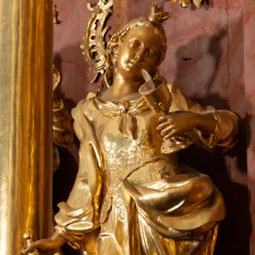 Zdjęcie nr 1: Rzeźba drążona, ustawiona na podstawie. Święta ukazana frontalnie, w kontrapoście, o esowato wygiętej sylwetce, z silnie wypchniętym lewym biodrem i głową przechyloną na lewe ramię. W lewej ugiętej i uniesionej ręce trzyma kielich z hostią, w prawej, opuszczonej miecz skierowany ostrzem w dół, na którym opiera ciężar ciała. Twarz ma owalną, o wydatnym podbródku, małych, migdałowatych oczach, prostym nosie i pełnych ustach. Włosy ma długie, kręcone, upięte z tyłu głowy, z pojedynczymi wijącymi się puklami opadającymi na lewe ramię, z wplecionymi perłami. Na głowie ma koronę otwartą. Ubrana jest w długą suknię, ściśle przylegającą do ciała, dekorowaną przy dekolcie szarfą, z gorsetem w talii zdobionym płaskorzeźbionym ornamentem roślinnym. Na prawe ramię ma narzucony płaszcz, opadający na lewy bok, zawiązany z przodu postaci. Figura w całości złocona, ostrze miecza i hostia srebrzone.