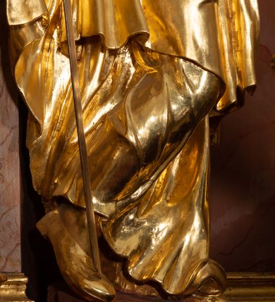 Zdjęcie nr 5: Rzeźba drążona, ustawiona na podstawie. Święta ukazana frontalnie, w kontrapoście, o esowato wygiętej sylwetce, z silnie wypchniętym lewym biodrem i głową przechyloną na lewe ramię. W lewej ugiętej i uniesionej ręce trzyma kielich z hostią, w prawej, opuszczonej miecz skierowany ostrzem w dół, na którym opiera ciężar ciała. Twarz ma owalną, o wydatnym podbródku, małych, migdałowatych oczach, prostym nosie i pełnych ustach. Włosy ma długie, kręcone, upięte z tyłu głowy, z pojedynczymi wijącymi się puklami opadającymi na lewe ramię, z wplecionymi perłami. Na głowie ma koronę otwartą. Ubrana jest w długą suknię, ściśle przylegającą do ciała, dekorowaną przy dekolcie szarfą, z gorsetem w talii zdobionym płaskorzeźbionym ornamentem roślinnym. Na prawe ramię ma narzucony płaszcz, opadający na lewy bok, zawiązany z przodu postaci. Figura w całości złocona, ostrze miecza i hostia srebrzone.