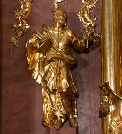 Zdjęcie nr 1: Rzeźba drążona, ustawiona na nieregularnej podstawie. Święty ukazany frontalnie, z głową skierowaną w lewo, w kontrapoście. Lewą rękę ma ugiętą i uniesioną w bok, w dłoni trzyma klucze, prawą ugiętą, przy boku, w której trzyma zamkniętą księgę. Twarz ma pociągłą, szczupłą, o małych, migdałowatych oczach, prostym nosie i pełnych, rozchylonych ustach, okoloną krótką, kręconą brodą i włosami. Ubrany jest w tunikę, rozchyloną na piersi, ściśle przylegająca do ciała, przepasaną w talii oraz obfity płaszcz zarzucony na prawe ramię i zawinięty na lewym boku i z przodu postaci. Figura w całości złocona.