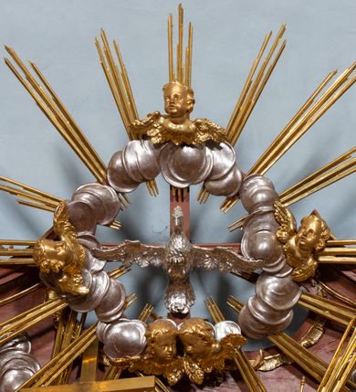Zdjęcie nr 3: W polu zwieńczenia grupa rzeźbiarska Trójcy Świętej, tronującej pośród obłoków, w glorii promienistej. 
Po lewej Chrystus w pozycji siedzącej, ukazany frontalnie, z głową przechyloną na lewe ramię, prawą ręką przytrzymuje z przodu krzyż, lewą kładzie na piersi. Twarz ma pociągłą, o migdałowatych oczach, krótkim nosie i rozchylonych ustach, okoloną krótką, kręconą brodą i kręconymi włosami, sięgającymi ramion. Ubrany jest w płaszcz zawinięty wokół dolnej partii ciała i na lewym ramieniu, odsłaniający klatkę piersiową. Bóg Ojciec po prawej stronie w pozycji siedzącej, zwrócony w trzech czwartych w prawo, z pochyloną głową, lewą rękę kładzie na piersi, w dłoni trzyma berło, prawą ugiętą wyciąga w bok. Twarz ma pociągłą, o migdałowatych oczach, krótkim nosie i rozchylonych ustach, okoloną kręconą brodą i włosami sięgającymi ramion. Wokół głowy ma trójkątny nimb. Ubrany jest w tunikę przepasaną w talii, przylegającą do ciała oraz płaszcz zawinięty wokół nóg i lewego ramienia. Pomiędzy postaciami na uskrzydlonej główce anielskiej unosi się glob. Nad nimi gołębica Ducha Świętego w otoku z obłoków i uskrzydlonych główek anielskich, w glorii promienistej. Postacie, aniołki i gloria złocone, obłoki, glob i gołębica srebrzone. 
