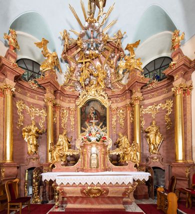 Zdjęcie nr 2: W polu zwieńczenia grupa rzeźbiarska Trójcy Świętej, tronującej pośród obłoków, w glorii promienistej. 
Po lewej Chrystus w pozycji siedzącej, ukazany frontalnie, z głową przechyloną na lewe ramię, prawą ręką przytrzymuje z przodu krzyż, lewą kładzie na piersi. Twarz ma pociągłą, o migdałowatych oczach, krótkim nosie i rozchylonych ustach, okoloną krótką, kręconą brodą i kręconymi włosami, sięgającymi ramion. Ubrany jest w płaszcz zawinięty wokół dolnej partii ciała i na lewym ramieniu, odsłaniający klatkę piersiową. Bóg Ojciec po prawej stronie w pozycji siedzącej, zwrócony w trzech czwartych w prawo, z pochyloną głową, lewą rękę kładzie na piersi, w dłoni trzyma berło, prawą ugiętą wyciąga w bok. Twarz ma pociągłą, o migdałowatych oczach, krótkim nosie i rozchylonych ustach, okoloną kręconą brodą i włosami sięgającymi ramion. Wokół głowy ma trójkątny nimb. Ubrany jest w tunikę przepasaną w talii, przylegającą do ciała oraz płaszcz zawinięty wokół nóg i lewego ramienia. Pomiędzy postaciami na uskrzydlonej główce anielskiej unosi się glob. Nad nimi gołębica Ducha Świętego w otoku z obłoków i uskrzydlonych główek anielskich, w glorii promienistej. Postacie, aniołki i gloria złocone, obłoki, glob i gołębica srebrzone. 
