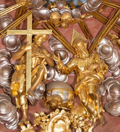 Zdjęcie nr 1: W polu zwieńczenia grupa rzeźbiarska Trójcy Świętej, tronującej pośród obłoków, w glorii promienistej. 
Po lewej Chrystus w pozycji siedzącej, ukazany frontalnie, z głową przechyloną na lewe ramię, prawą ręką przytrzymuje z przodu krzyż, lewą kładzie na piersi. Twarz ma pociągłą, o migdałowatych oczach, krótkim nosie i rozchylonych ustach, okoloną krótką, kręconą brodą i kręconymi włosami, sięgającymi ramion. Ubrany jest w płaszcz zawinięty wokół dolnej partii ciała i na lewym ramieniu, odsłaniający klatkę piersiową. Bóg Ojciec po prawej stronie w pozycji siedzącej, zwrócony w trzech czwartych w prawo, z pochyloną głową, lewą rękę kładzie na piersi, w dłoni trzyma berło, prawą ugiętą wyciąga w bok. Twarz ma pociągłą, o migdałowatych oczach, krótkim nosie i rozchylonych ustach, okoloną kręconą brodą i włosami sięgającymi ramion. Wokół głowy ma trójkątny nimb. Ubrany jest w tunikę przepasaną w talii, przylegającą do ciała oraz płaszcz zawinięty wokół nóg i lewego ramienia. Pomiędzy postaciami na uskrzydlonej główce anielskiej unosi się glob. Nad nimi gołębica Ducha Świętego w otoku z obłoków i uskrzydlonych główek anielskich, w glorii promienistej. Postacie, aniołki i gloria złocone, obłoki, glob i gołębica srebrzone. 
