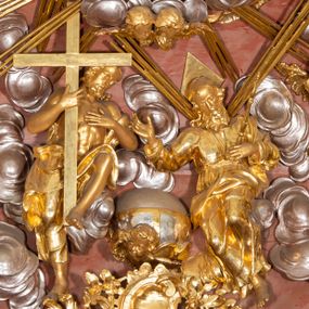 Zdjęcie nr 1: W polu zwieńczenia grupa rzeźbiarska Trójcy Świętej, tronującej pośród obłoków, w glorii promienistej. 
Po lewej Chrystus w pozycji siedzącej, ukazany frontalnie, z głową przechyloną na lewe ramię, prawą ręką przytrzymuje z przodu krzyż, lewą kładzie na piersi. Twarz ma pociągłą, o migdałowatych oczach, krótkim nosie i rozchylonych ustach, okoloną krótką, kręconą brodą i kręconymi włosami, sięgającymi ramion. Ubrany jest w płaszcz zawinięty wokół dolnej partii ciała i na lewym ramieniu, odsłaniający klatkę piersiową. Bóg Ojciec po prawej stronie w pozycji siedzącej, zwrócony w trzech czwartych w prawo, z pochyloną głową, lewą rękę kładzie na piersi, w dłoni trzyma berło, prawą ugiętą wyciąga w bok. Twarz ma pociągłą, o migdałowatych oczach, krótkim nosie i rozchylonych ustach, okoloną kręconą brodą i włosami sięgającymi ramion. Wokół głowy ma trójkątny nimb. Ubrany jest w tunikę przepasaną w talii, przylegającą do ciała oraz płaszcz zawinięty wokół nóg i lewego ramienia. Pomiędzy postaciami na uskrzydlonej główce anielskiej unosi się glob. Nad nimi gołębica Ducha Świętego w otoku z obłoków i uskrzydlonych główek anielskich, w glorii promienistej. Postacie, aniołki i gloria złocone, obłoki, glob i gołębica srebrzone. 
