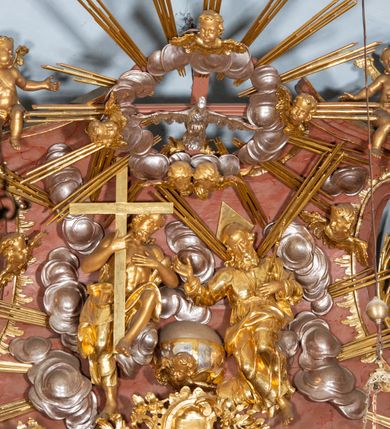 Zdjęcie nr 6: Nastawa ołtarzowa architektoniczna, jednokondygnacyjna ze zwieńczeniem, pięcioosiowa, ustawiona na wysokim cokole. Ołtarz wolnostojący, drewniany, sarkofagowy, wklęsło-wypukły, o zdwojonym froncie; mensa drewniana z marmurowym portatylem. Od frontu rocaille'owy kartusz ujęty fantazyjnymi płycinami oraz liśćmi. Na ołtarzu tabernakulum w formie sześcianu o wklęsłych bokach i wklęsło-wypukłym froncie; ujęte w narożach dwiema parami pilastrów o trzonach przechodzących w spływy wolutowe zdobione rocaille'm i różami. Pilastry podtrzymują falujący gzyms oraz fragmenty falistego, przerwanego, odwróconego przyczółka, przechodzącego w cokół, stanowiący tron wystawienia, na którym stoi krucyfiks. Na ściance frontowej obrotowa nisza, o wykroju stojącego prostokąta zamkniętego łukiem półkolistym, w opasce, zwieńczona kampanulami. W jej polu drzwiczki w formie stojącego prostokąta zamkniętego łukiem półkolistym w profilowanej ramie, z odlewanym przedstawieniem baranka leżącego na księdze z pieczęciami. Tabernakulum ujęte spływami przechodzącymi na krańcach we fragmenty wolutowego gzymsu, na których siedzą figury adorujących aniołów. Ponad tabernakulum fantazyjna ścianka tworząca tło dla tronu wystawienia z prześwitem otoczonym obłokami, z glorią promienistą, flankowana rocaille'owymi uszami. Cokół nastawy z wyodrębnionymi dwoma prostopadłościanami i dwoma konsolami, na dwóch skrajnych osiach bramki zamknięte wolutowymi szczytami tworzącymi cokoły dla figur, z przejściami w formie stojącego prostokąta zamkniętego łukiem odcinkowym nadwieszonym, z drzwiczkami. Cokół i drzwiczki dekorowane rocaille'm. Lico nastawy wklęsłe, osie wydzielone trzema parami pilastrów, na których tle stoją dwie pary kolumn o kapitelach kompozytowych. Podpory dźwigają pełne belkowanie, wyłamane na ich osi, przerwane nad polem środkowym, o gzymsie przechodzącym miejscami w rocaille'owe kartusze. Na osi środkowej wnęka z obrazem Matki Boskiej Wspomożenia Wiernych, w formie stojącego prostokąta zamkniętego łukiem odcinkowym nadwieszonym z uskokiem, ujęta profilowaną ramą, dekorowaną ponad łukiem płomienistym rocaille'm. Na pozostałych czterech osiach figury św. Barbary, św. Apolonii, św. Piotra i św. Pawła ustawione na rokokowych wspornikach. Trzony pilastrów i pola osi bocznych wypełnione rocaille'm, fryz dekorowany zwisami kwiatowymi. Zwieńczenie w formie zbliżonej do trapezu, zamknięte dwoma fragmentami gzymsu przechodzącego w woluty, flankowane wolutami spływającymi na belkowanie wewnętrznej pary kolumn, na których siedzą adorujące figury aniołów. W polu rzeźbiarska grupa Trójcy Świętej, pośród obłoków, w glorii promienistej, zwieńczona gołębicą Ducha Świętego w otoku z obłoków i uskrzydlonych główek anielskich, w glorii promienistej, flankowana przez dwa putta siedzące na gzymsie. Zwieńczenie po bokach obwiedzione rocaille'm, ujęte liśćmi palmowymi. Na belkowaniu, na osiach zewnętrznej pary kolumn wolutowe cokoły z rokokowymi wazonami. Struktura polichromowana na sposób imitujący marmoryzację w kolorze różowym i różowo-pomarańczowym, kolumny, profile i ornamentyka złocone, obłoki srebrzone. 

