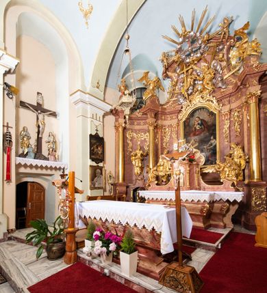 Zdjęcie nr 4: Nastawa ołtarzowa architektoniczna, jednokondygnacyjna ze zwieńczeniem, pięcioosiowa, ustawiona na wysokim cokole. Ołtarz wolnostojący, drewniany, sarkofagowy, wklęsło-wypukły, o zdwojonym froncie; mensa drewniana z marmurowym portatylem. Od frontu rocaille'owy kartusz ujęty fantazyjnymi płycinami oraz liśćmi. Na ołtarzu tabernakulum w formie sześcianu o wklęsłych bokach i wklęsło-wypukłym froncie; ujęte w narożach dwiema parami pilastrów o trzonach przechodzących w spływy wolutowe zdobione rocaille'm i różami. Pilastry podtrzymują falujący gzyms oraz fragmenty falistego, przerwanego, odwróconego przyczółka, przechodzącego w cokół, stanowiący tron wystawienia, na którym stoi krucyfiks. Na ściance frontowej obrotowa nisza, o wykroju stojącego prostokąta zamkniętego łukiem półkolistym, w opasce, zwieńczona kampanulami. W jej polu drzwiczki w formie stojącego prostokąta zamkniętego łukiem półkolistym w profilowanej ramie, z odlewanym przedstawieniem baranka leżącego na księdze z pieczęciami. Tabernakulum ujęte spływami przechodzącymi na krańcach we fragmenty wolutowego gzymsu, na których siedzą figury adorujących aniołów. Ponad tabernakulum fantazyjna ścianka tworząca tło dla tronu wystawienia z prześwitem otoczonym obłokami, z glorią promienistą, flankowana rocaille'owymi uszami. Cokół nastawy z wyodrębnionymi dwoma prostopadłościanami i dwoma konsolami, na dwóch skrajnych osiach bramki zamknięte wolutowymi szczytami tworzącymi cokoły dla figur, z przejściami w formie stojącego prostokąta zamkniętego łukiem odcinkowym nadwieszonym, z drzwiczkami. Cokół i drzwiczki dekorowane rocaille'm. Lico nastawy wklęsłe, osie wydzielone trzema parami pilastrów, na których tle stoją dwie pary kolumn o kapitelach kompozytowych. Podpory dźwigają pełne belkowanie, wyłamane na ich osi, przerwane nad polem środkowym, o gzymsie przechodzącym miejscami w rocaille'owe kartusze. Na osi środkowej wnęka z obrazem Matki Boskiej Wspomożenia Wiernych, w formie stojącego prostokąta zamkniętego łukiem odcinkowym nadwieszonym z uskokiem, ujęta profilowaną ramą, dekorowaną ponad łukiem płomienistym rocaille'm. Na pozostałych czterech osiach figury św. Barbary, św. Apolonii, św. Piotra i św. Pawła ustawione na rokokowych wspornikach. Trzony pilastrów i pola osi bocznych wypełnione rocaille'm, fryz dekorowany zwisami kwiatowymi. Zwieńczenie w formie zbliżonej do trapezu, zamknięte dwoma fragmentami gzymsu przechodzącego w woluty, flankowane wolutami spływającymi na belkowanie wewnętrznej pary kolumn, na których siedzą adorujące figury aniołów. W polu rzeźbiarska grupa Trójcy Świętej, pośród obłoków, w glorii promienistej, zwieńczona gołębicą Ducha Świętego w otoku z obłoków i uskrzydlonych główek anielskich, w glorii promienistej, flankowana przez dwa putta siedzące na gzymsie. Zwieńczenie po bokach obwiedzione rocaille'm, ujęte liśćmi palmowymi. Na belkowaniu, na osiach zewnętrznej pary kolumn wolutowe cokoły z rokokowymi wazonami. Struktura polichromowana na sposób imitujący marmoryzację w kolorze różowym i różowo-pomarańczowym, kolumny, profile i ornamentyka złocone, obłoki srebrzone. 

