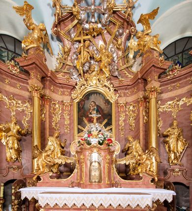 Zdjęcie nr 3: Nastawa ołtarzowa architektoniczna, jednokondygnacyjna ze zwieńczeniem, pięcioosiowa, ustawiona na wysokim cokole. Ołtarz wolnostojący, drewniany, sarkofagowy, wklęsło-wypukły, o zdwojonym froncie; mensa drewniana z marmurowym portatylem. Od frontu rocaille'owy kartusz ujęty fantazyjnymi płycinami oraz liśćmi. Na ołtarzu tabernakulum w formie sześcianu o wklęsłych bokach i wklęsło-wypukłym froncie; ujęte w narożach dwiema parami pilastrów o trzonach przechodzących w spływy wolutowe zdobione rocaille'm i różami. Pilastry podtrzymują falujący gzyms oraz fragmenty falistego, przerwanego, odwróconego przyczółka, przechodzącego w cokół, stanowiący tron wystawienia, na którym stoi krucyfiks. Na ściance frontowej obrotowa nisza, o wykroju stojącego prostokąta zamkniętego łukiem półkolistym, w opasce, zwieńczona kampanulami. W jej polu drzwiczki w formie stojącego prostokąta zamkniętego łukiem półkolistym w profilowanej ramie, z odlewanym przedstawieniem baranka leżącego na księdze z pieczęciami. Tabernakulum ujęte spływami przechodzącymi na krańcach we fragmenty wolutowego gzymsu, na których siedzą figury adorujących aniołów. Ponad tabernakulum fantazyjna ścianka tworząca tło dla tronu wystawienia z prześwitem otoczonym obłokami, z glorią promienistą, flankowana rocaille'owymi uszami. Cokół nastawy z wyodrębnionymi dwoma prostopadłościanami i dwoma konsolami, na dwóch skrajnych osiach bramki zamknięte wolutowymi szczytami tworzącymi cokoły dla figur, z przejściami w formie stojącego prostokąta zamkniętego łukiem odcinkowym nadwieszonym, z drzwiczkami. Cokół i drzwiczki dekorowane rocaille'm. Lico nastawy wklęsłe, osie wydzielone trzema parami pilastrów, na których tle stoją dwie pary kolumn o kapitelach kompozytowych. Podpory dźwigają pełne belkowanie, wyłamane na ich osi, przerwane nad polem środkowym, o gzymsie przechodzącym miejscami w rocaille'owe kartusze. Na osi środkowej wnęka z obrazem Matki Boskiej Wspomożenia Wiernych, w formie stojącego prostokąta zamkniętego łukiem odcinkowym nadwieszonym z uskokiem, ujęta profilowaną ramą, dekorowaną ponad łukiem płomienistym rocaille'm. Na pozostałych czterech osiach figury św. Barbary, św. Apolonii, św. Piotra i św. Pawła ustawione na rokokowych wspornikach. Trzony pilastrów i pola osi bocznych wypełnione rocaille'm, fryz dekorowany zwisami kwiatowymi. Zwieńczenie w formie zbliżonej do trapezu, zamknięte dwoma fragmentami gzymsu przechodzącego w woluty, flankowane wolutami spływającymi na belkowanie wewnętrznej pary kolumn, na których siedzą adorujące figury aniołów. W polu rzeźbiarska grupa Trójcy Świętej, pośród obłoków, w glorii promienistej, zwieńczona gołębicą Ducha Świętego w otoku z obłoków i uskrzydlonych główek anielskich, w glorii promienistej, flankowana przez dwa putta siedzące na gzymsie. Zwieńczenie po bokach obwiedzione rocaille'm, ujęte liśćmi palmowymi. Na belkowaniu, na osiach zewnętrznej pary kolumn wolutowe cokoły z rokokowymi wazonami. Struktura polichromowana na sposób imitujący marmoryzację w kolorze różowym i różowo-pomarańczowym, kolumny, profile i ornamentyka złocone, obłoki srebrzone. 

