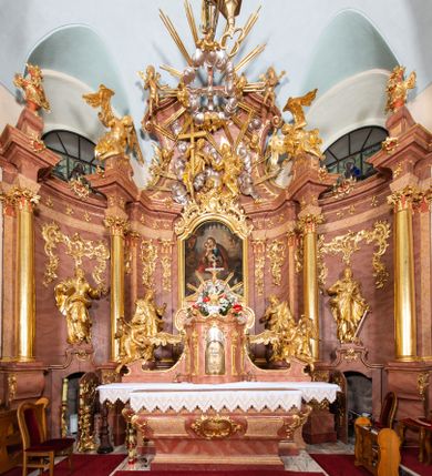 Zdjęcie nr 1: Nastawa ołtarzowa architektoniczna, jednokondygnacyjna ze zwieńczeniem, pięcioosiowa, ustawiona na wysokim cokole. Ołtarz wolnostojący, drewniany, sarkofagowy, wklęsło-wypukły, o zdwojonym froncie; mensa drewniana z marmurowym portatylem. Od frontu rocaille'owy kartusz ujęty fantazyjnymi płycinami oraz liśćmi. Na ołtarzu tabernakulum w formie sześcianu o wklęsłych bokach i wklęsło-wypukłym froncie; ujęte w narożach dwiema parami pilastrów o trzonach przechodzących w spływy wolutowe zdobione rocaille'm i różami. Pilastry podtrzymują falujący gzyms oraz fragmenty falistego, przerwanego, odwróconego przyczółka, przechodzącego w cokół, stanowiący tron wystawienia, na którym stoi krucyfiks. Na ściance frontowej obrotowa nisza, o wykroju stojącego prostokąta zamkniętego łukiem półkolistym, w opasce, zwieńczona kampanulami. W jej polu drzwiczki w formie stojącego prostokąta zamkniętego łukiem półkolistym w profilowanej ramie, z odlewanym przedstawieniem baranka leżącego na księdze z pieczęciami. Tabernakulum ujęte spływami przechodzącymi na krańcach we fragmenty wolutowego gzymsu, na których siedzą figury adorujących aniołów. Ponad tabernakulum fantazyjna ścianka tworząca tło dla tronu wystawienia z prześwitem otoczonym obłokami, z glorią promienistą, flankowana rocaille'owymi uszami. Cokół nastawy z wyodrębnionymi dwoma prostopadłościanami i dwoma konsolami, na dwóch skrajnych osiach bramki zamknięte wolutowymi szczytami tworzącymi cokoły dla figur, z przejściami w formie stojącego prostokąta zamkniętego łukiem odcinkowym nadwieszonym, z drzwiczkami. Cokół i drzwiczki dekorowane rocaille'm. Lico nastawy wklęsłe, osie wydzielone trzema parami pilastrów, na których tle stoją dwie pary kolumn o kapitelach kompozytowych. Podpory dźwigają pełne belkowanie, wyłamane na ich osi, przerwane nad polem środkowym, o gzymsie przechodzącym miejscami w rocaille'owe kartusze. Na osi środkowej wnęka z obrazem Matki Boskiej Wspomożenia Wiernych, w formie stojącego prostokąta zamkniętego łukiem odcinkowym nadwieszonym z uskokiem, ujęta profilowaną ramą, dekorowaną ponad łukiem płomienistym rocaille'm. Na pozostałych czterech osiach figury św. Barbary, św. Apolonii, św. Piotra i św. Pawła ustawione na rokokowych wspornikach. Trzony pilastrów i pola osi bocznych wypełnione rocaille'm, fryz dekorowany zwisami kwiatowymi. Zwieńczenie w formie zbliżonej do trapezu, zamknięte dwoma fragmentami gzymsu przechodzącego w woluty, flankowane wolutami spływającymi na belkowanie wewnętrznej pary kolumn, na których siedzą adorujące figury aniołów. W polu rzeźbiarska grupa Trójcy Świętej, pośród obłoków, w glorii promienistej, zwieńczona gołębicą Ducha Świętego w otoku z obłoków i uskrzydlonych główek anielskich, w glorii promienistej, flankowana przez dwa putta siedzące na gzymsie. Zwieńczenie po bokach obwiedzione rocaille'm, ujęte liśćmi palmowymi. Na belkowaniu, na osiach zewnętrznej pary kolumn wolutowe cokoły z rokokowymi wazonami. Struktura polichromowana na sposób imitujący marmoryzację w kolorze różowym i różowo-pomarańczowym, kolumny, profile i ornamentyka złocone, obłoki srebrzone. 

